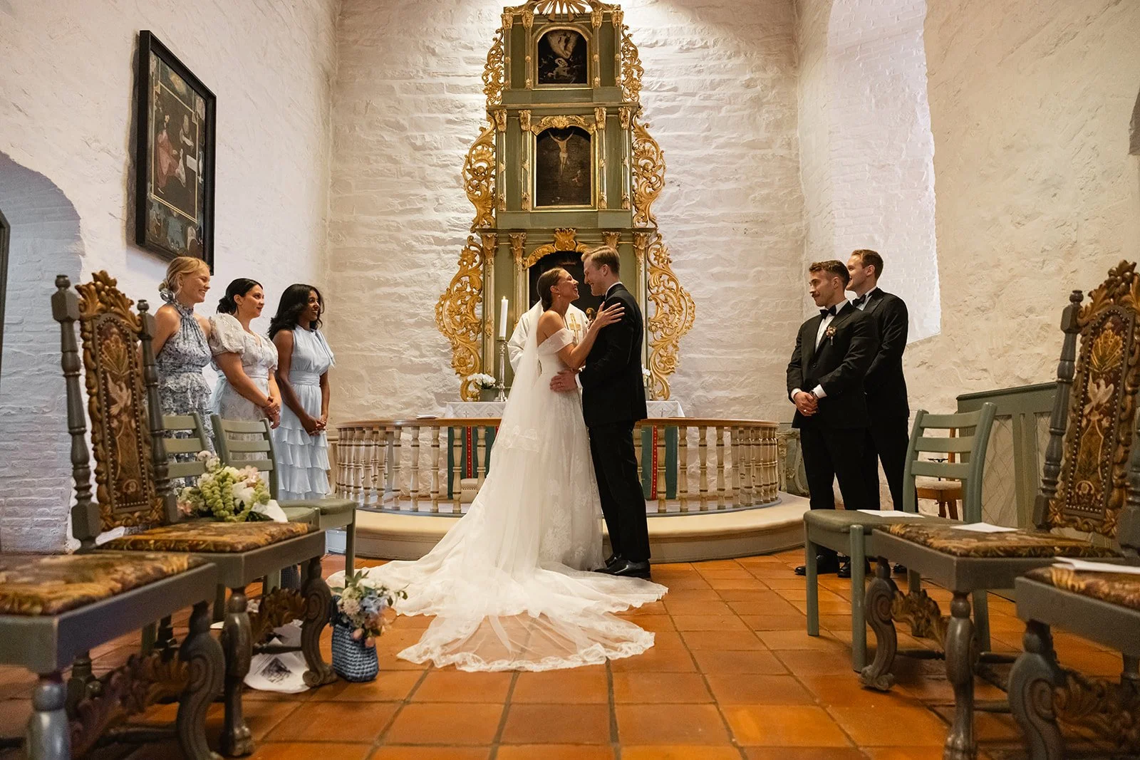 En brud og en brudgom står overfor hverandre i en kirke, holder hverandre i hendene, mens en presten eller offisiell sannsynligvis leder vielsen. To menn i dress står til høyre, og tre kvinner i kjoler står til venstre i kirken. Det er blomster og tr