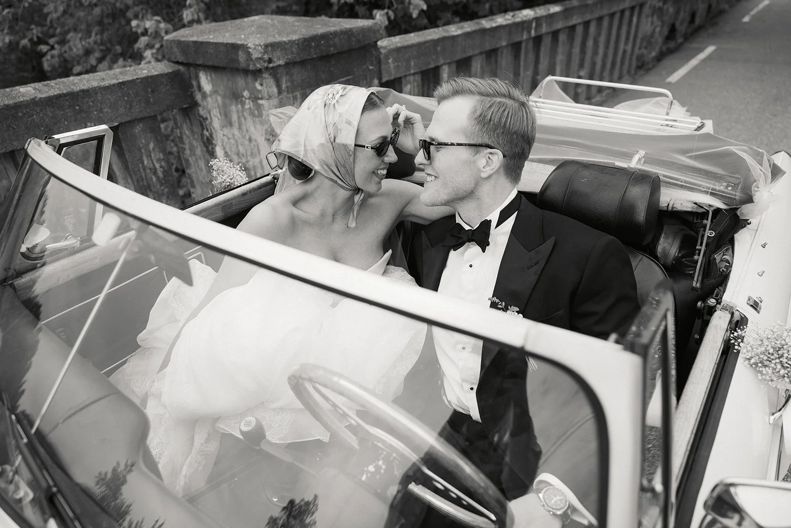 Et nygift ektepar kjører i en åpen bil på bryllupsdagen, begge smilende og nær hverandre.
