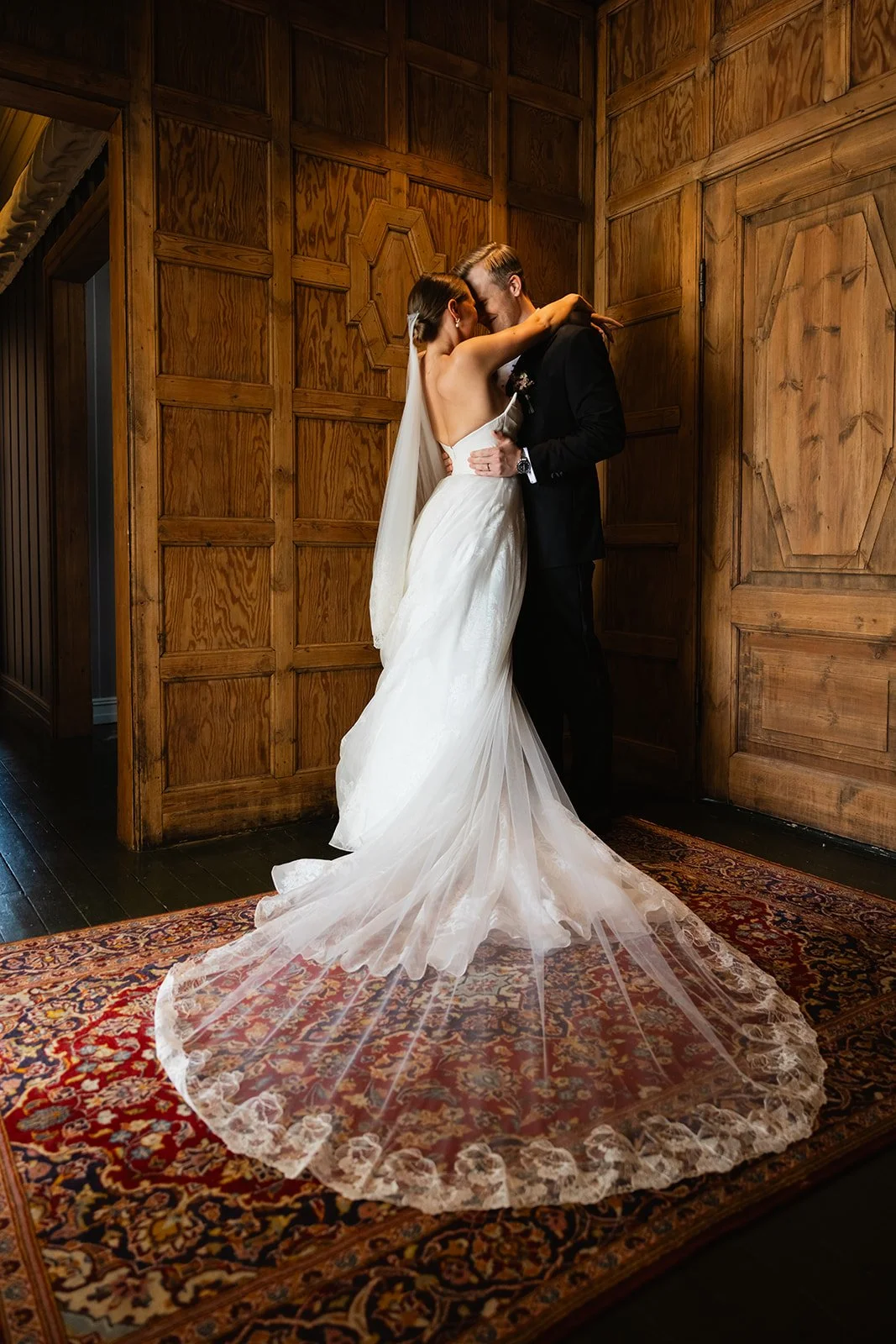Et brudepar i en omfavnelse står på en teppebelagt gulv inne i et rom med trepaneler på veggene.