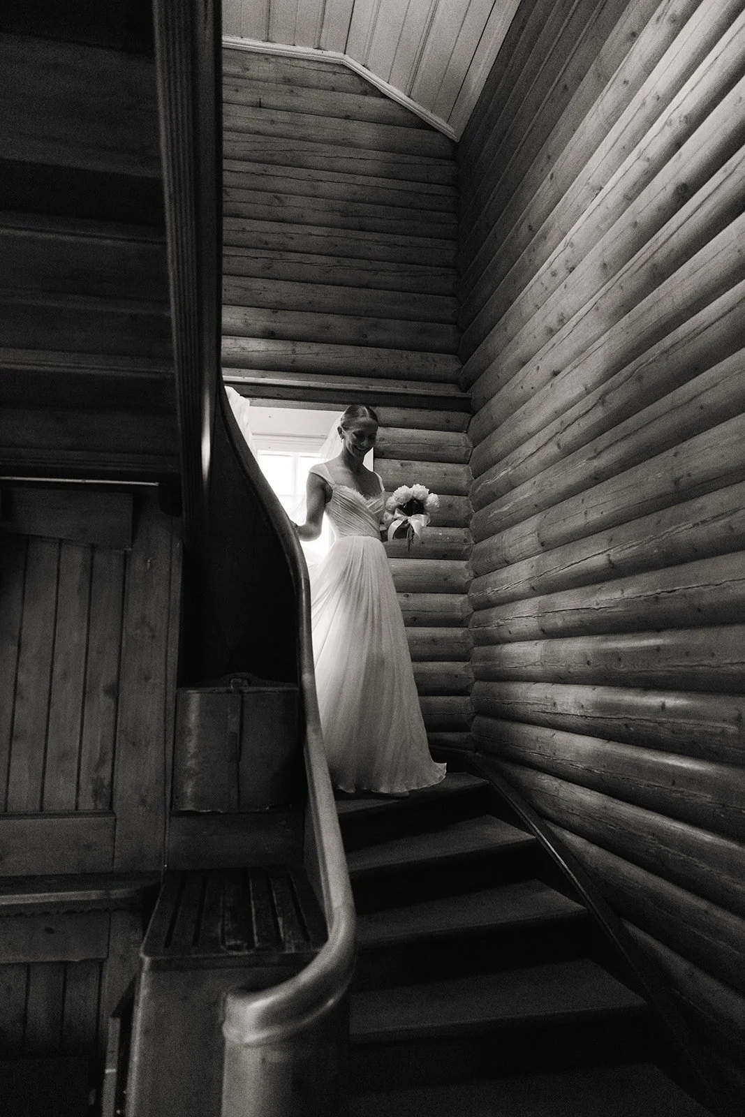 En kvinne i en brudekjole står på en trapp i et rom med tre vegger, tatt i svart-hvitt.
