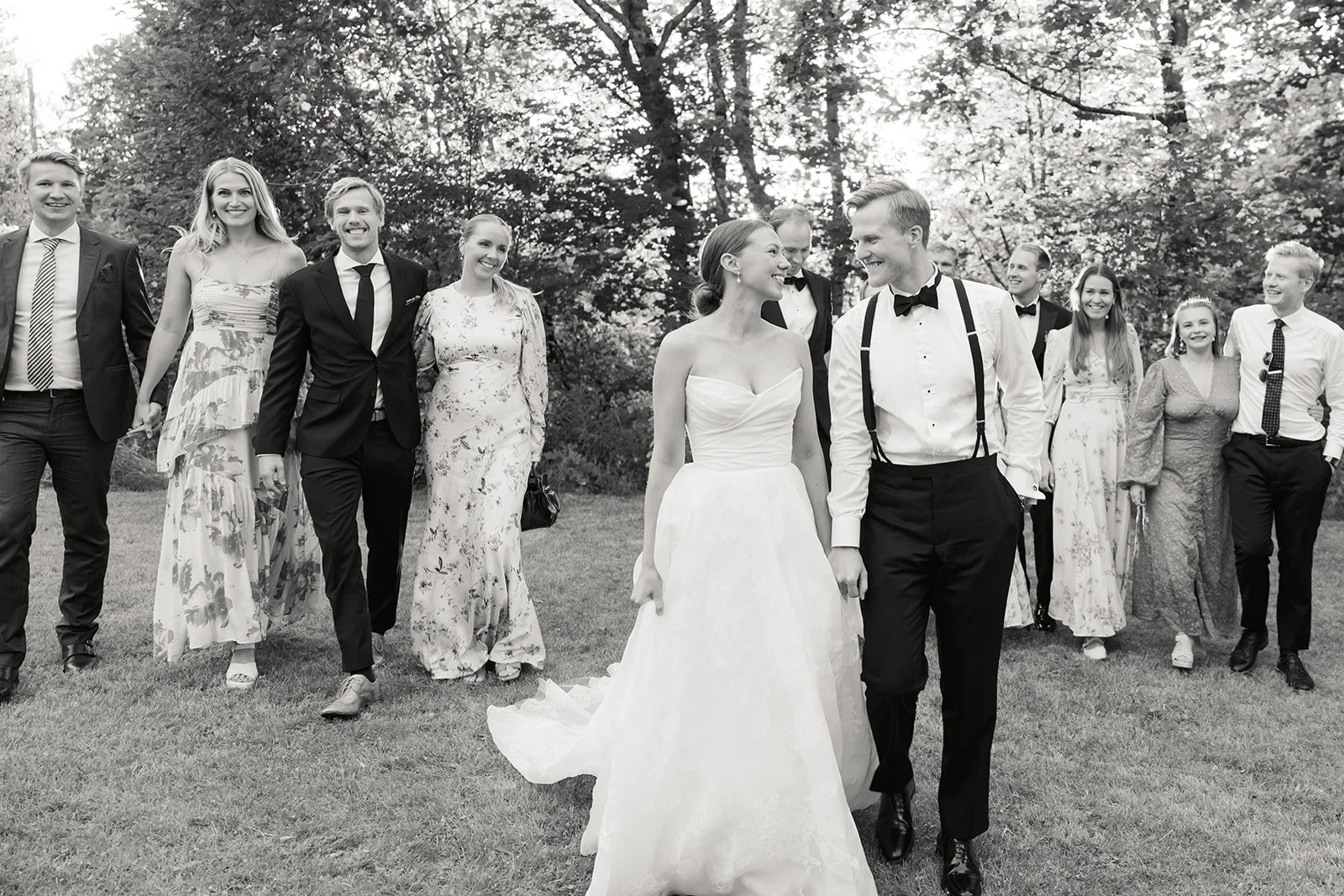 Et svart-hvitt bilde av et bryllup, der en brud og en brudgom går hånd i hånd igjen, omgitt av venner og familie som smiler og ler.