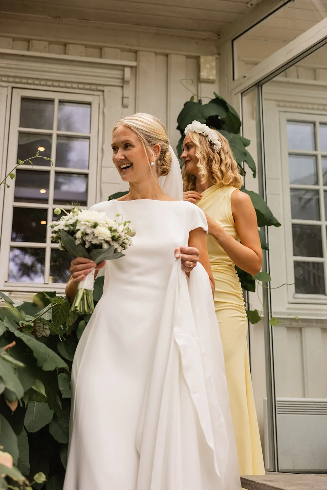 En brud i hvit bryllupsdress holder en blomsterbukett, smiler, og står i et rom med store vinduer og grønne planter, mens en kvinnelig gjest i lysegul kjole står bak henne, smilende og med blomsterkrans i håret.
