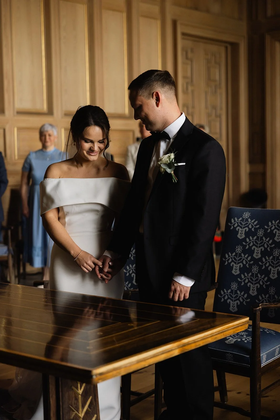 En brud og en brudgom står og holder hender under ekteskapsinngåelse, medvitne og andre i bakgrunnen i et litografisk seremonirom.