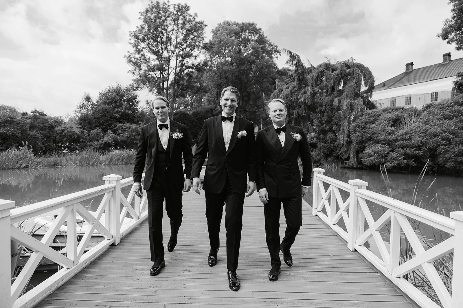 Tre menn i svarte dresser og smoking går på en trebro over vann, omgitt av trær og en bygning i bakgrunnen, svart-hvitt.