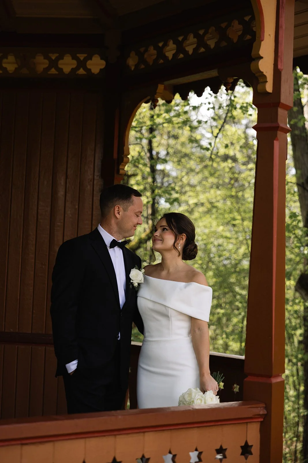 Et nygift par står nær hverandre på en liten trapp utendørs, med grønne trær i bakgrunnen, under en lystig, detaljert trekonstruksjon.