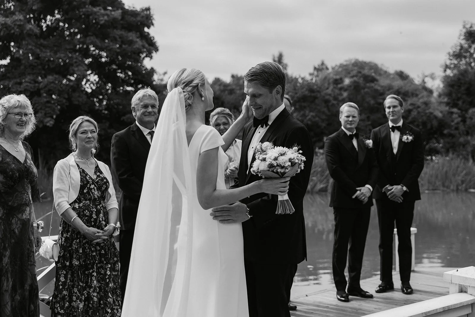 Et svart og hvitt bilde av et bryllups scena ved en sjø, der en brud og en brudgom står ansikt til ansikt. Bruden har på seg en lang kjole med rygg og en brudekåpe med slør, og holder en blomsterbukett. Brudgommen har på seg smoking med sløyfe. Flere