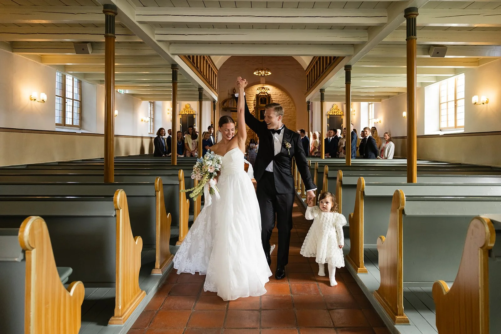 Nygift par i bryllupsklær som går ut av kirken, med en liten jente i hvit kjole, på en kirke med mennesker i bakgrunnen.