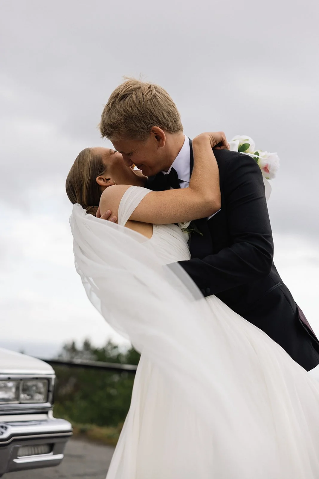 Et brudepar klemmer utendørs under en grå sky, hvor kvinnen i en hvit kjole holdes av en mann i dress.