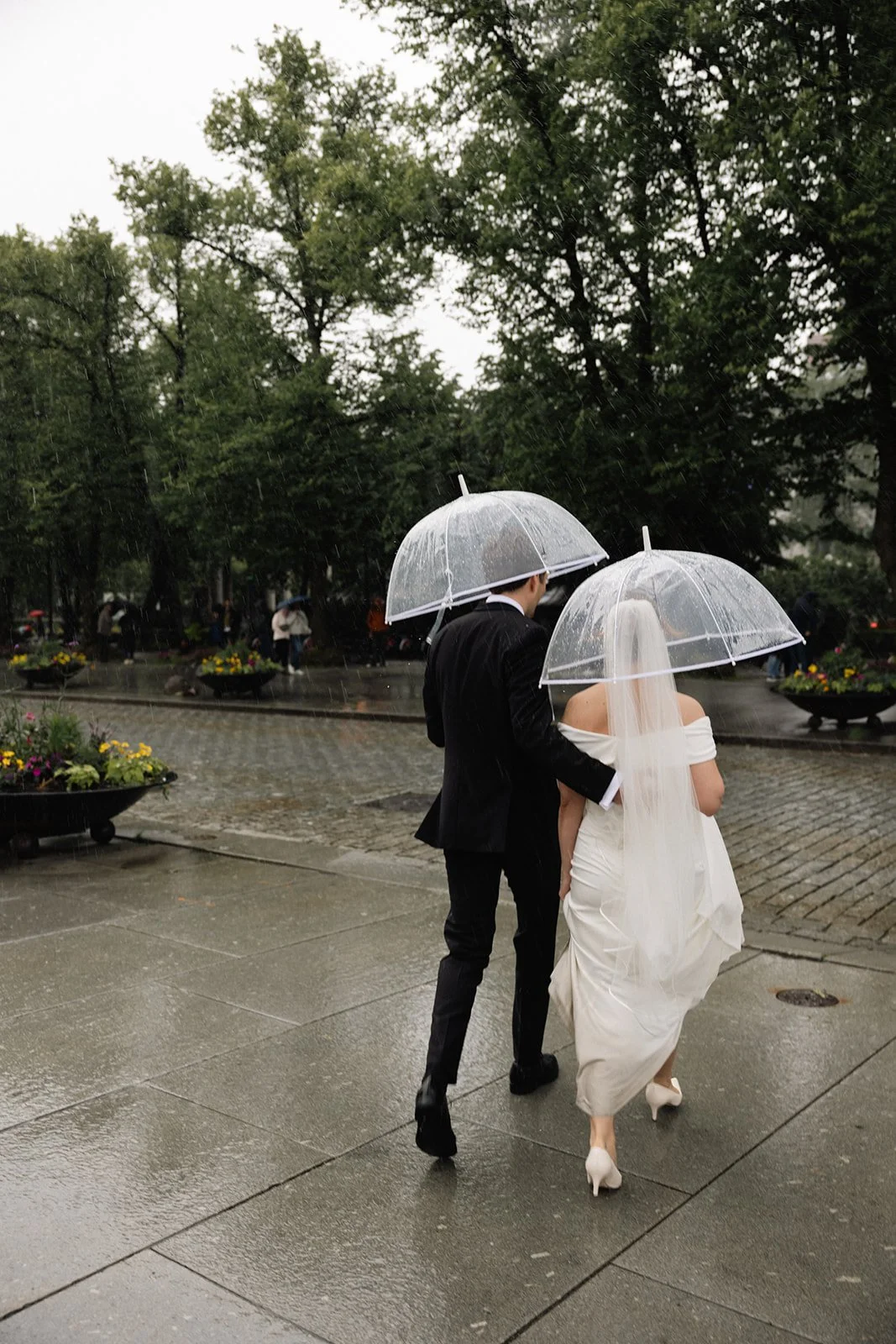 Et brudepar går hånd i hånd i regnet, med klare paraplyer, på en grå bygate med blomsterbed og trær.