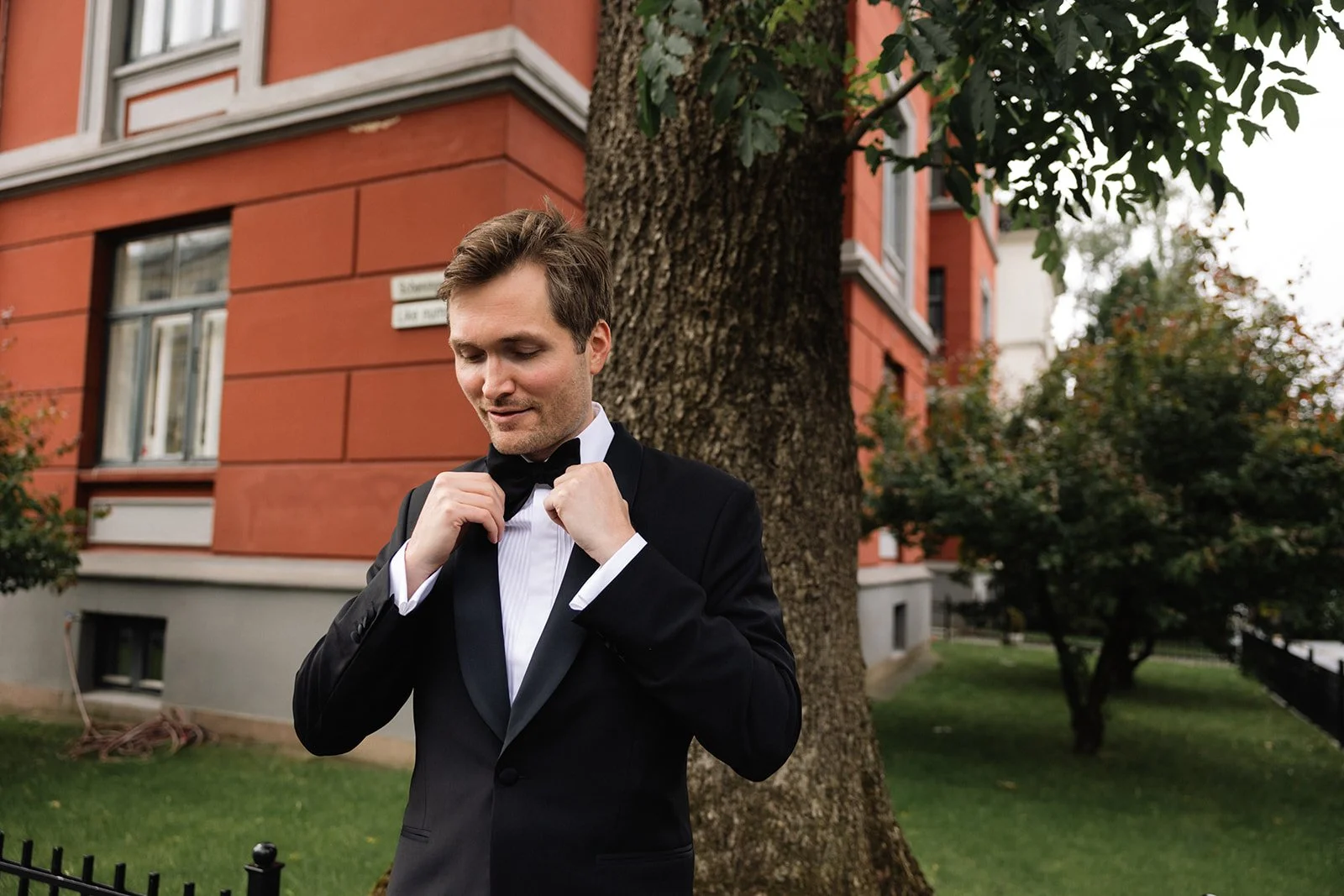 En mann i svart dress og sløyfe justerer sin slips foran et tre utenfor en bygning med oransje vegger.