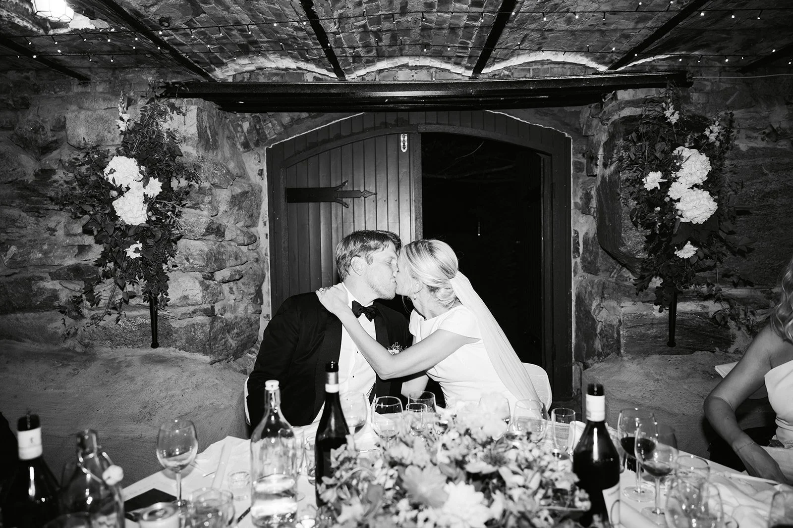 Et ektepar kysser under en bryllupsfest, omgitt av blomster og vinflasker, i et rom med steinmur og trerammer.