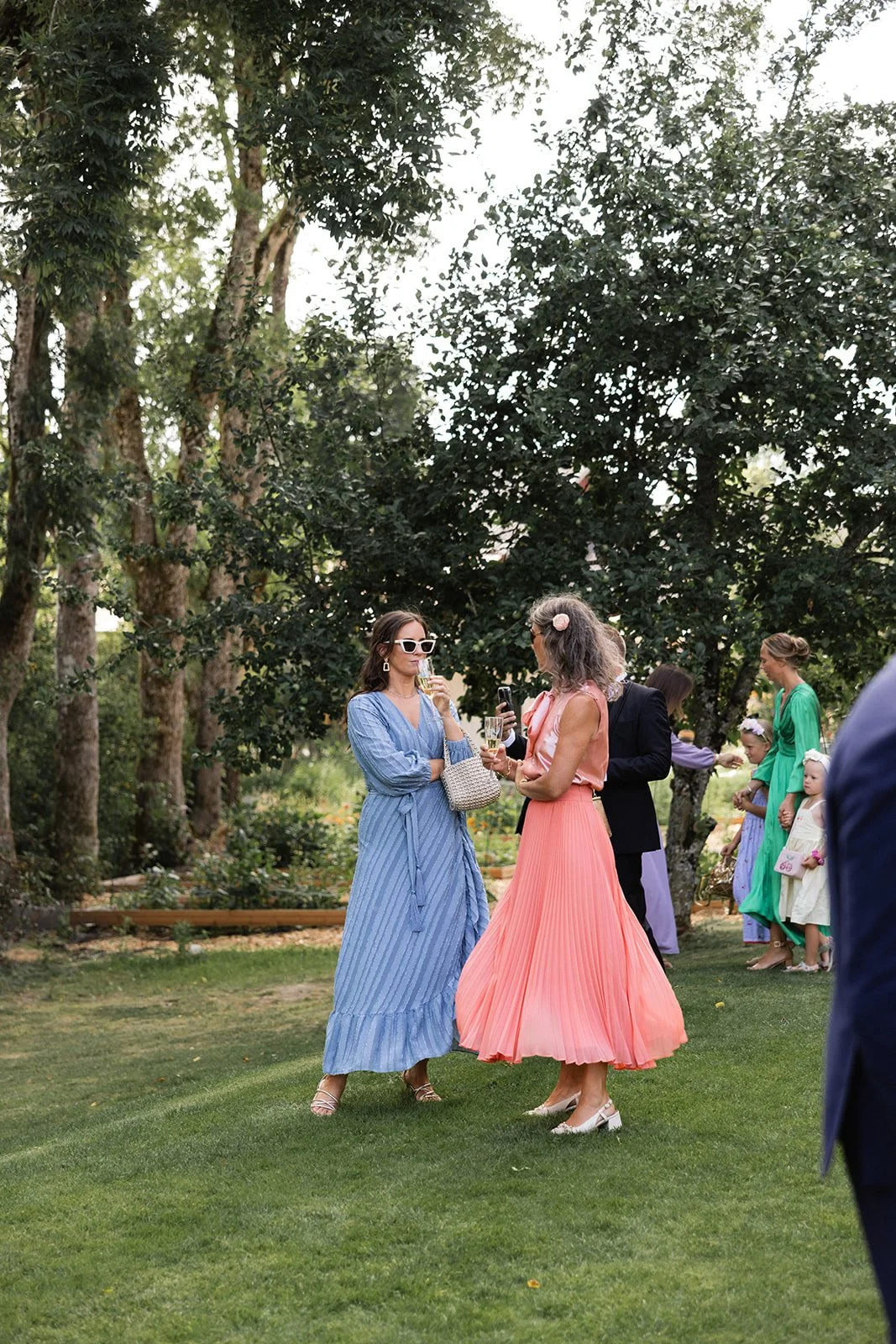 To kvinner i fargerike kjoler snakker og har drikke på en hagefest, med andre personer i kjole i bakgrunnen, omgitt av grønne trær.