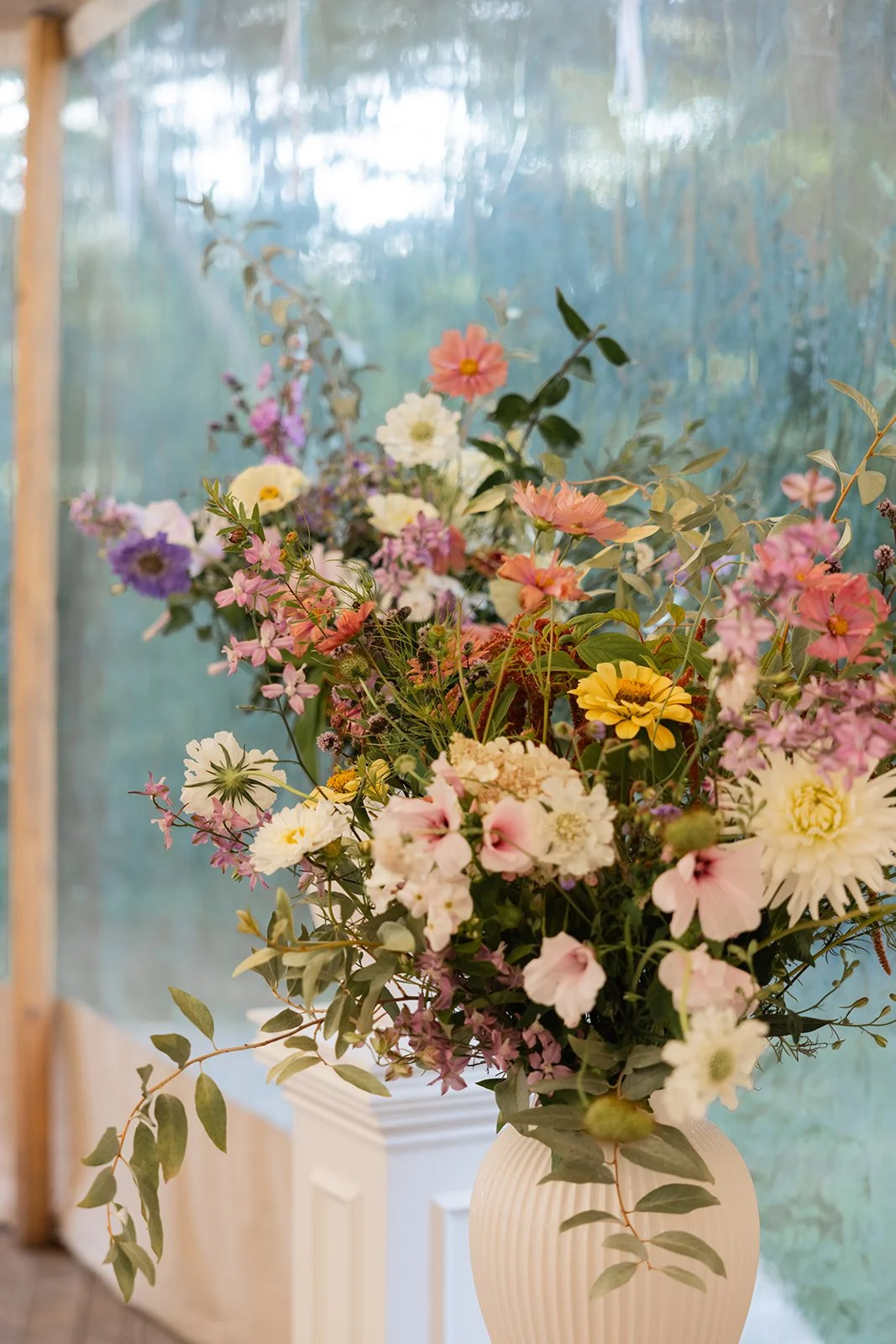 En stor vase med et fargerikt blomsterarrangement av ulike blomster og grønt, plassert på en hvit plint.
