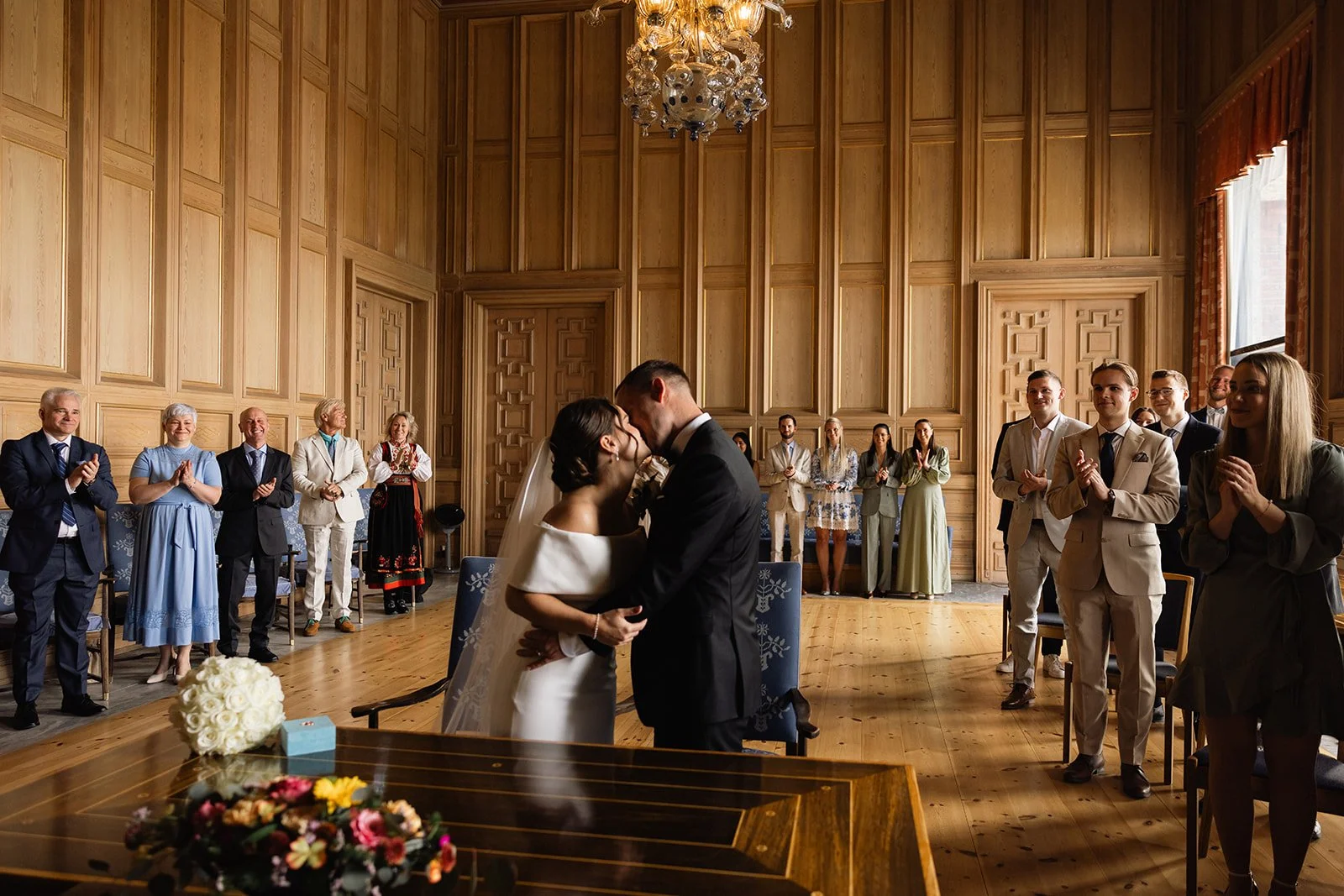 Et brudepar kysser i midten av en stor, treveggstaket rom med store vinduer og gardiner, omgitt av gjester som applauderer.