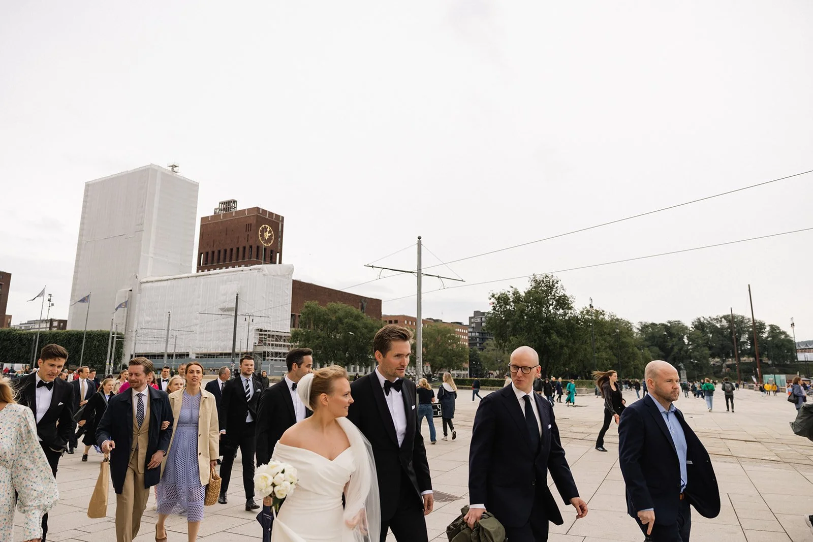 En gruppe mennesker, inkludert en brud med en hvit kjole og blomsterbukett, og en brudgom i svart dress, går på et utendørs område i en by