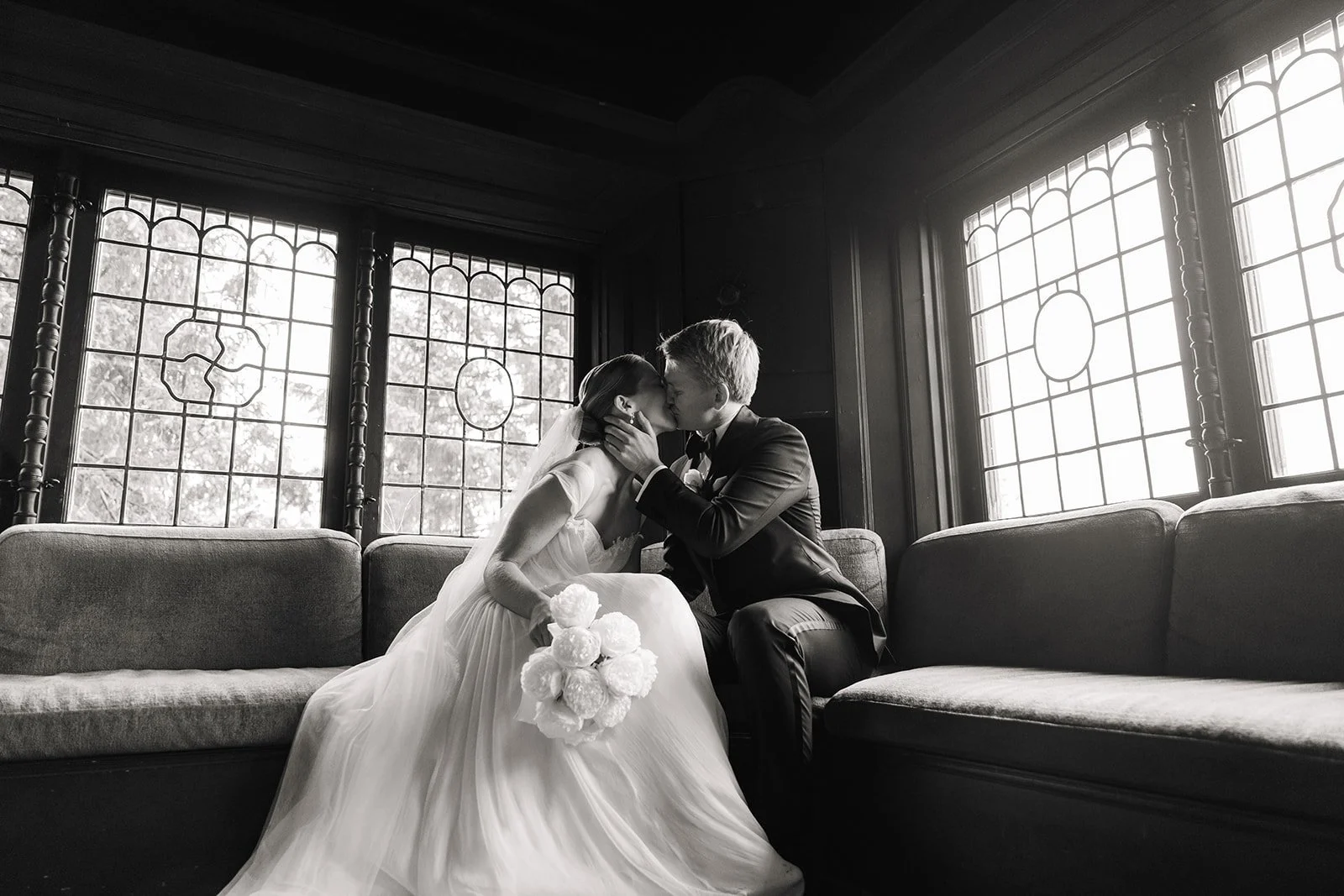 Et svart-hvitt bilde av et nygift par som kysser, mannen holder kvinnen i ansiktet, de sitter på en sofa i et rom med store vinduer med dekorative rammer.