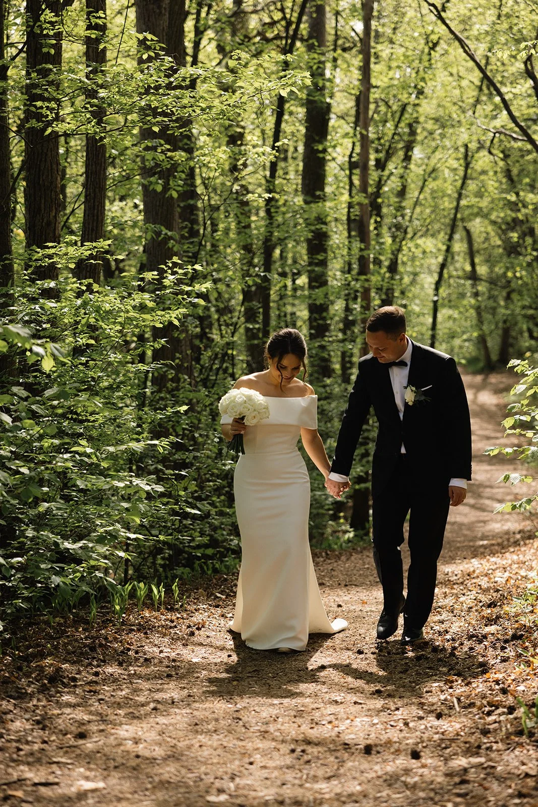 Et nygift par i brudekjole og mørk dress går hånd i hånd langs en skogssti.
