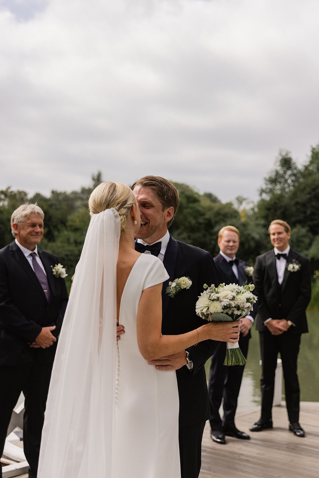 Et brudepar kysser under bryllupsseremoni med brudgom i smoking og brud i hvit kjole med brudekrage, omgitt av tre menn i svarte dresser og smykker, ved en innsjø utendørs.