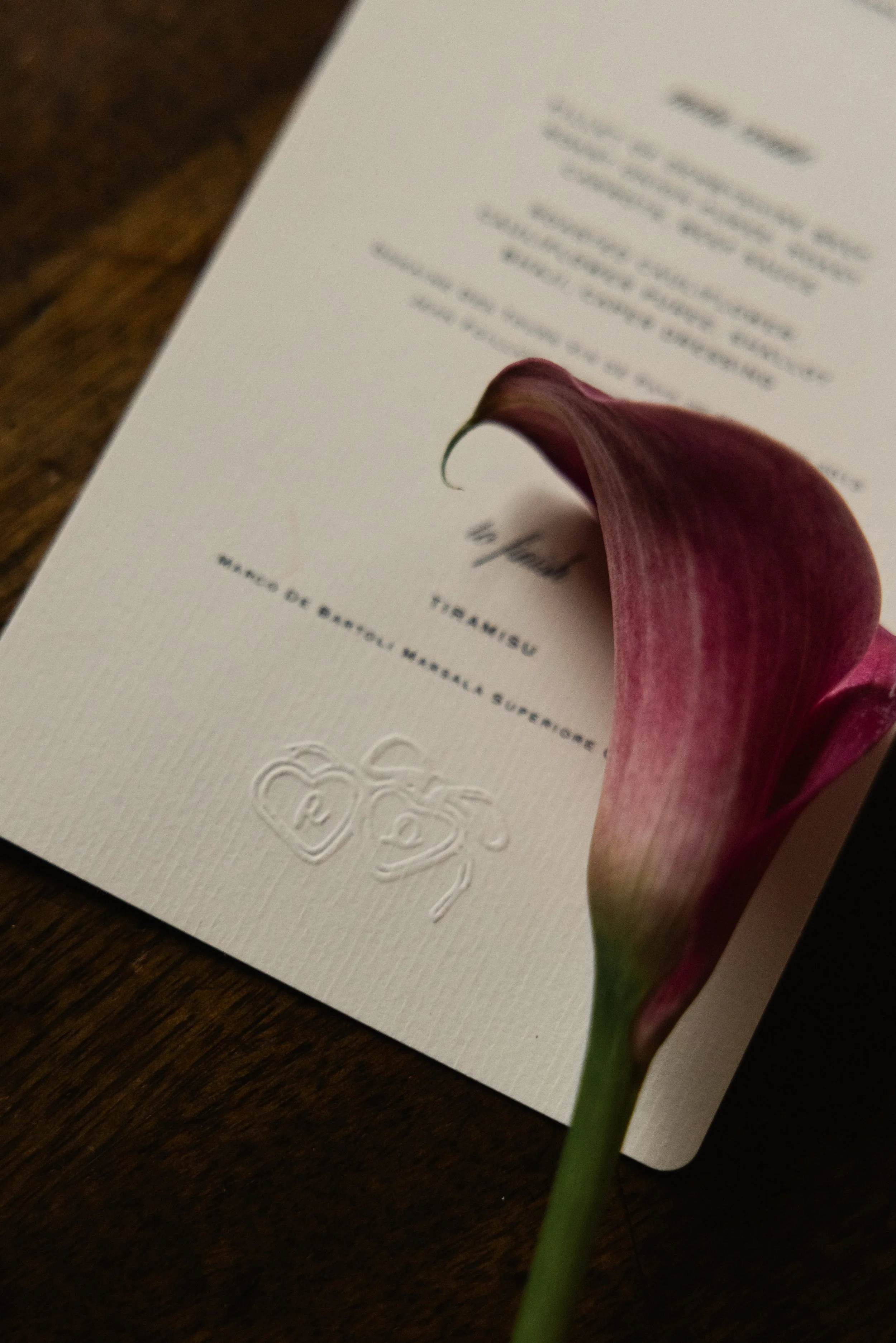 Et invitasjonskort med blomster, sannsynligvis en calla lily, ligger på en treflate.