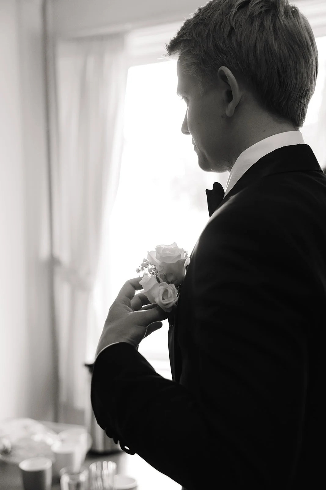 En mann i svart smoking og med sløyfe, holder en liten bukett av hvite roser, foran et vindu med lyse gardiner. Svart-hvitt bilde.