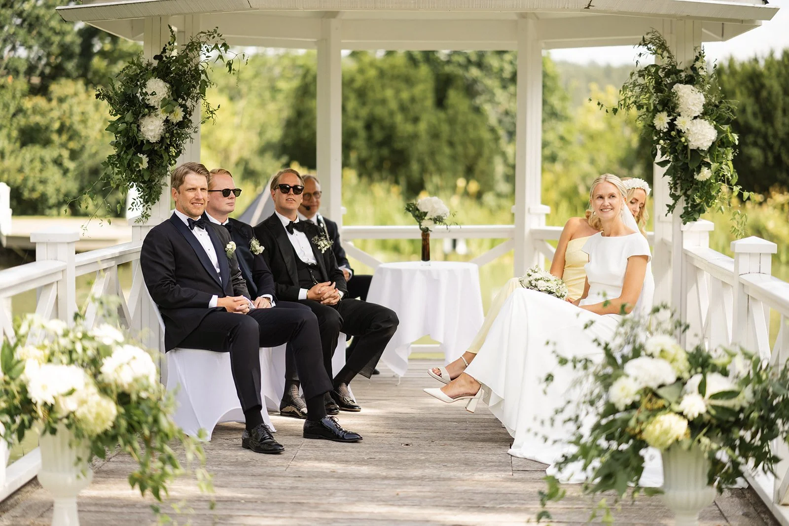 Et bryllupsscene med bruden og brudgommen som sitter på en plattform dekorert med hvite blomster, omgitt av gartnere og gjester i formelle klær, under en lystak serta med en grønn hage i bakgrunnen.