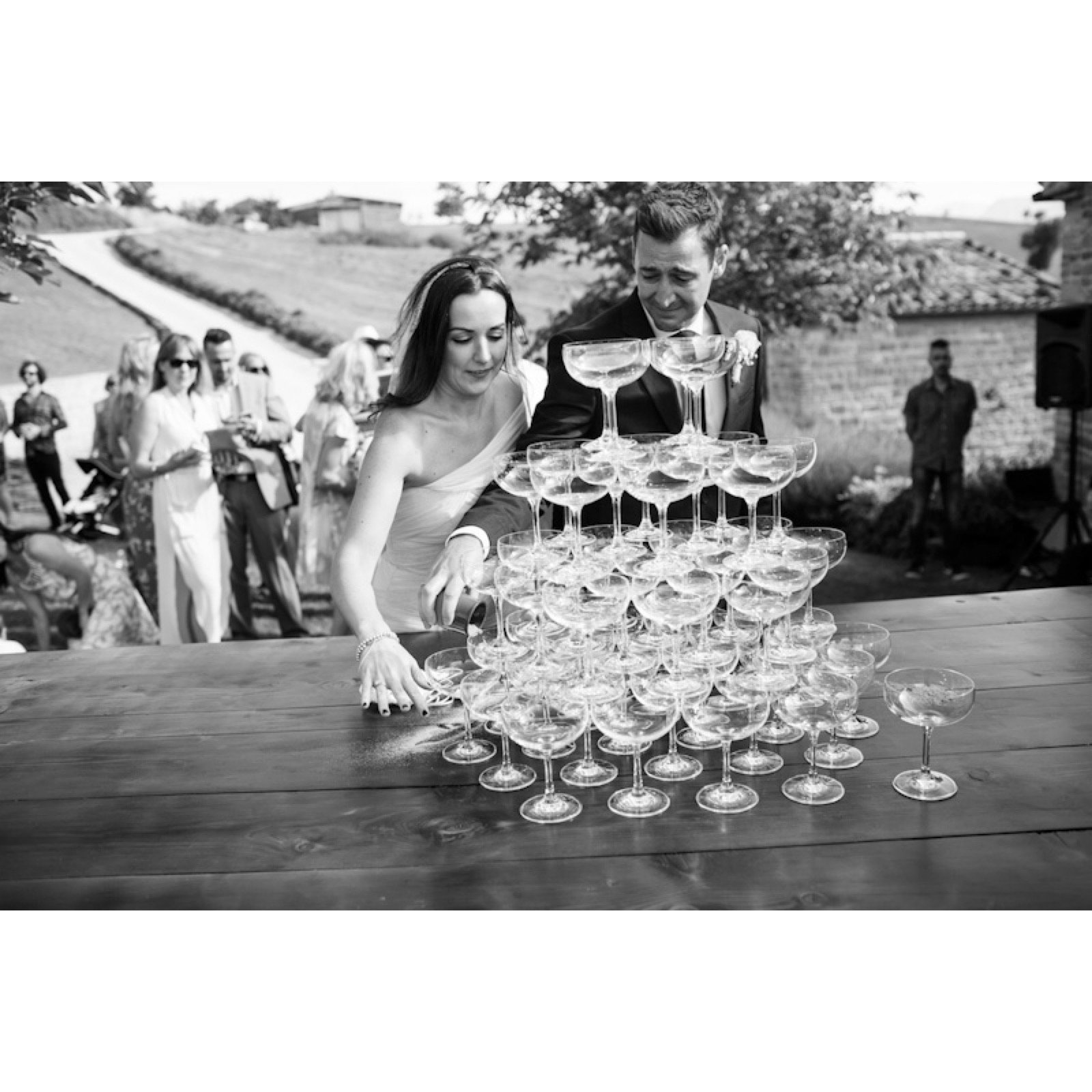A wedding celebration with a couple pouring champagne into a pyramid of glasses outdoors.