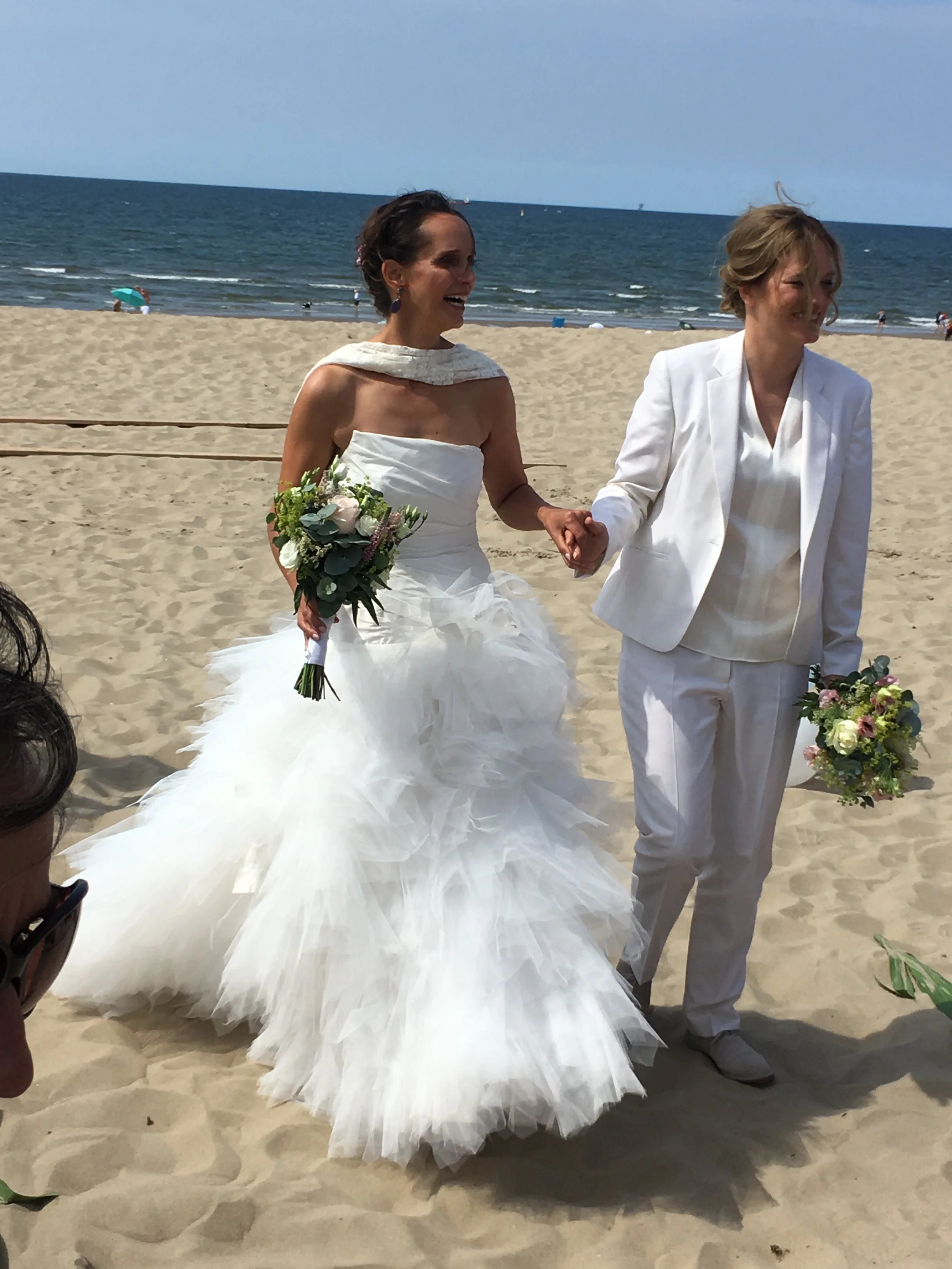 A bride and a person in a white suit holding hands on a sandy beach during a wedding ceremony, with the ocean and a blue sky in the background.