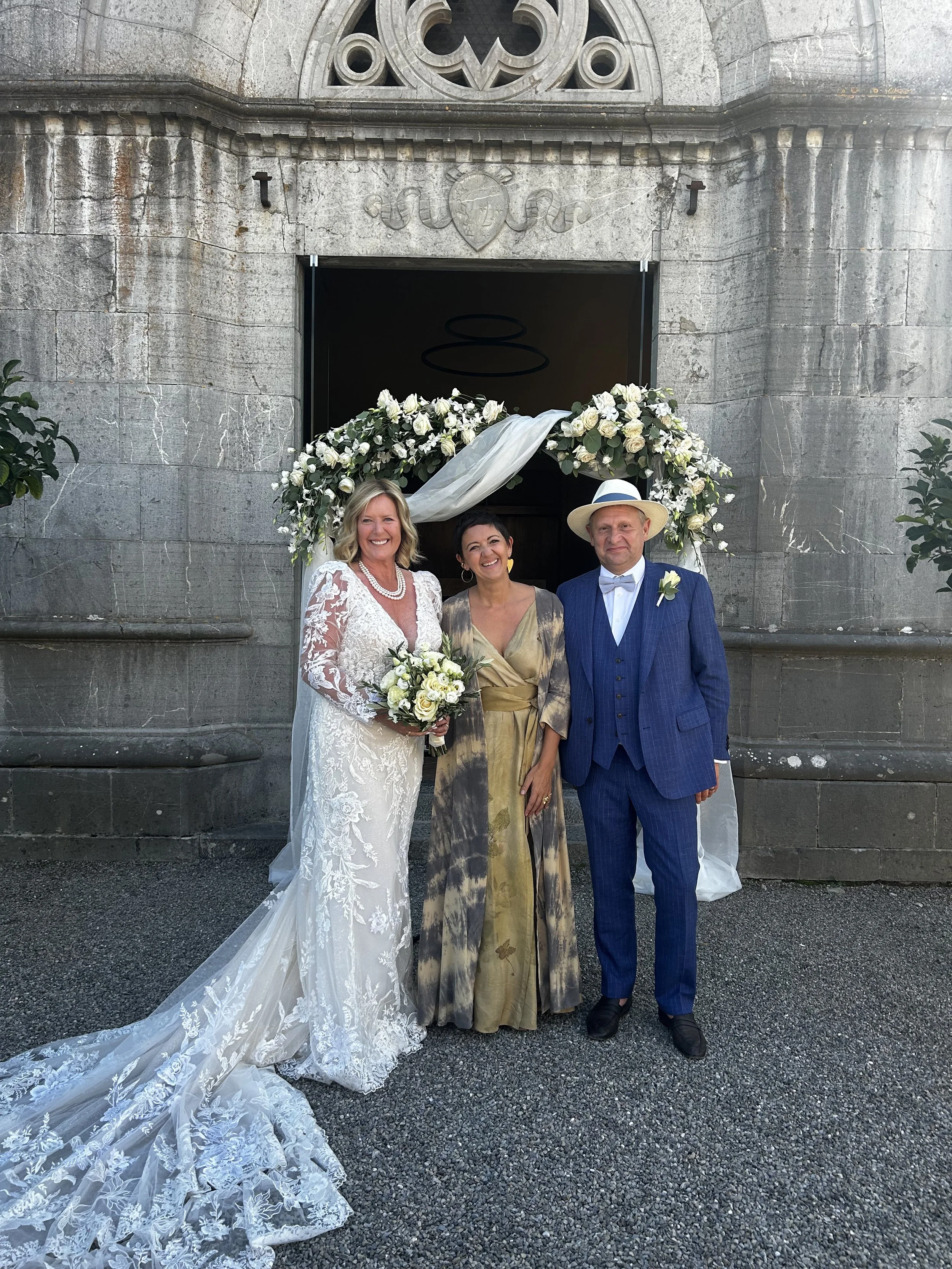 Three people standing in front of a wedding arch outside a stone building. The woman on the left is in a white lace wedding dress holding a bouquet. The woman in the middle is wearing a gold and green dress. The man on the right is in a blue suit with a white shirt and tie, and a straw hat with a blue band.