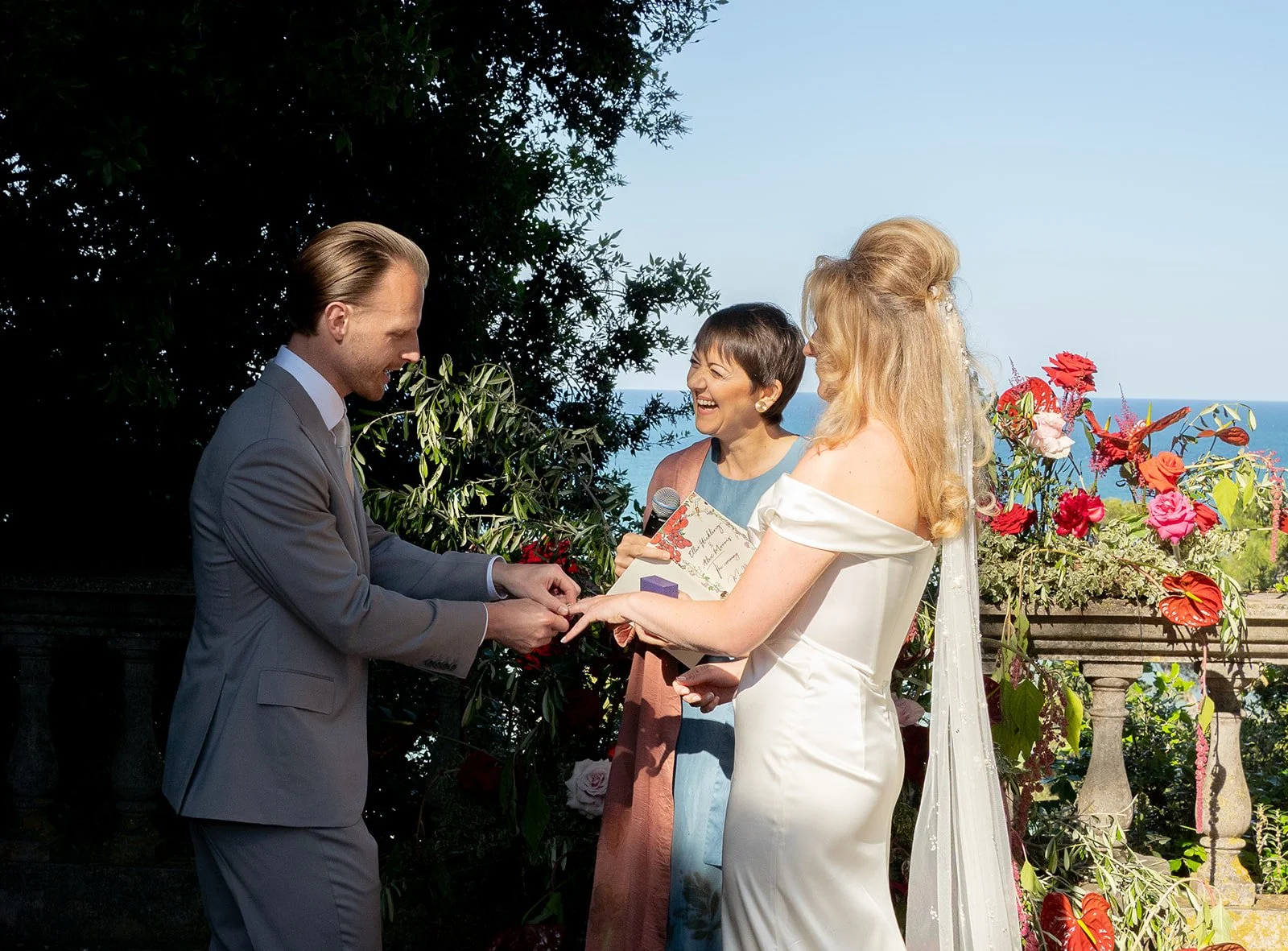 A wedding ceremony taking place outdoors with a bride, groom, and officiant. The groom is placing a ring on the bride's finger. There are flowers and greenery in the background, with a view of the ocean and sky.