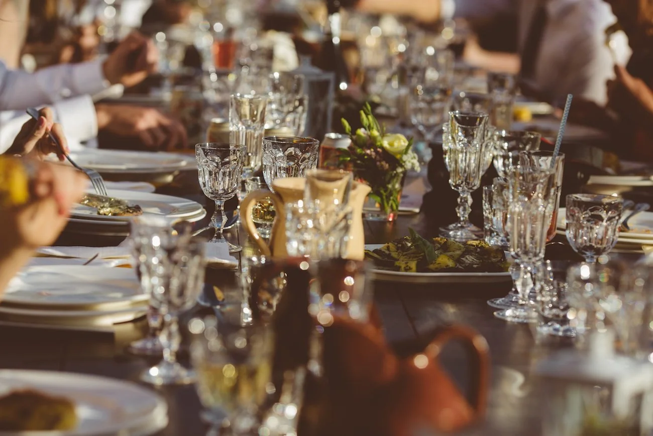 A long dining table set with glassware, plates, and a floral centerpiece, likely at an outdoor event or gathering.