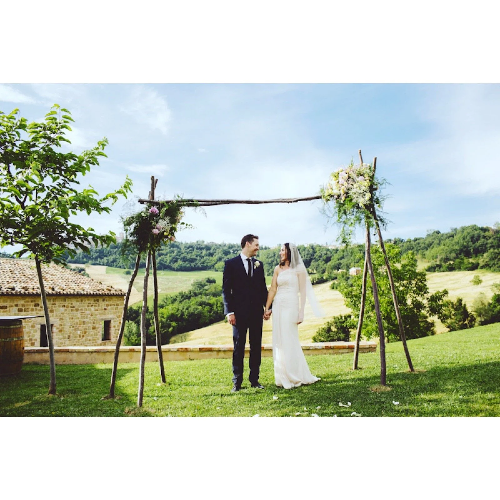 Bride and groom holding hands under a rustic wooden wedding arch with flowers, outdoors on a sunny day with green hills in the background.