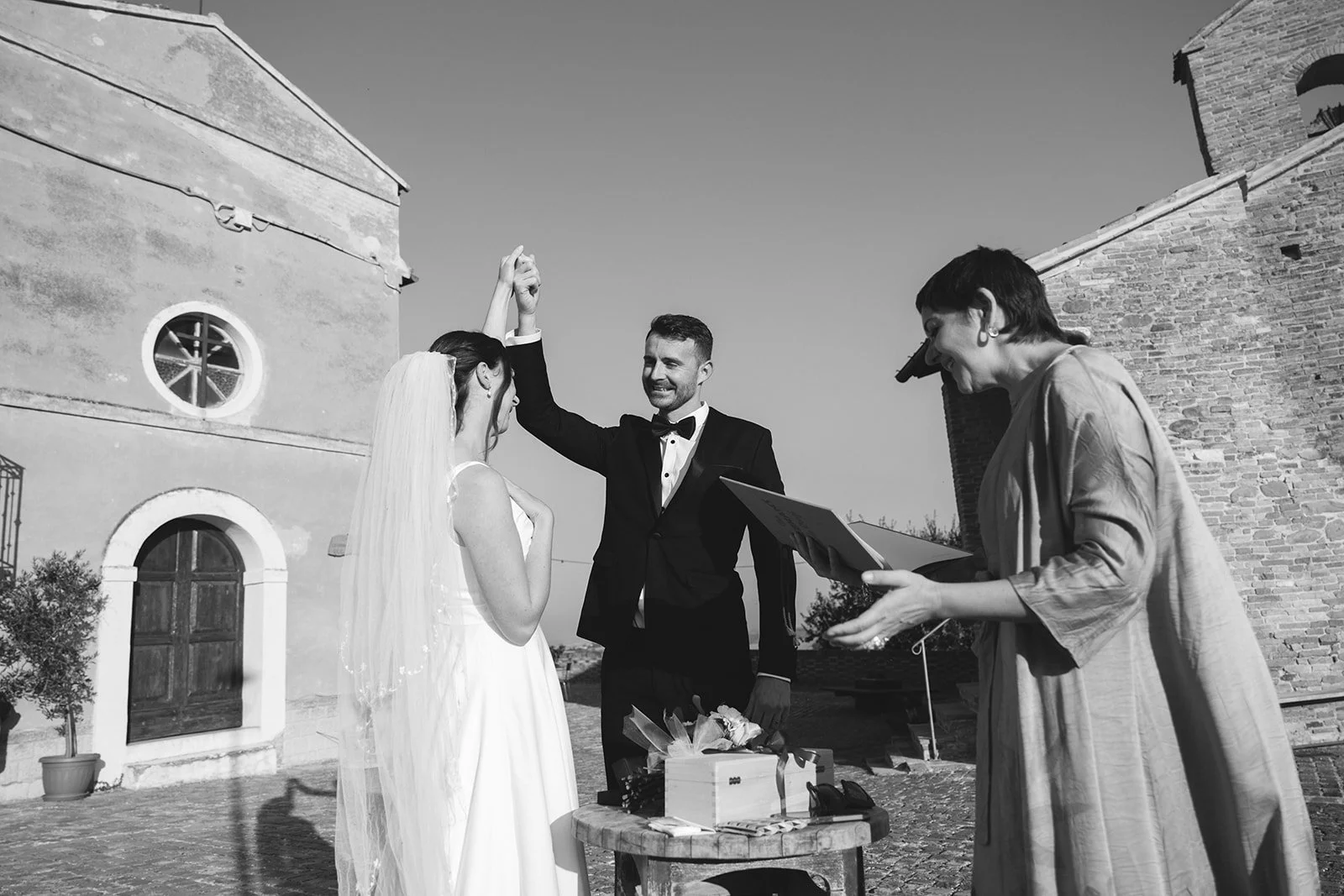 A bride and groom at their outdoor wedding ceremony with an officiant holding a tablet, as the groom raises his hand in celebration, in front of old brick buildings on a sunny day.