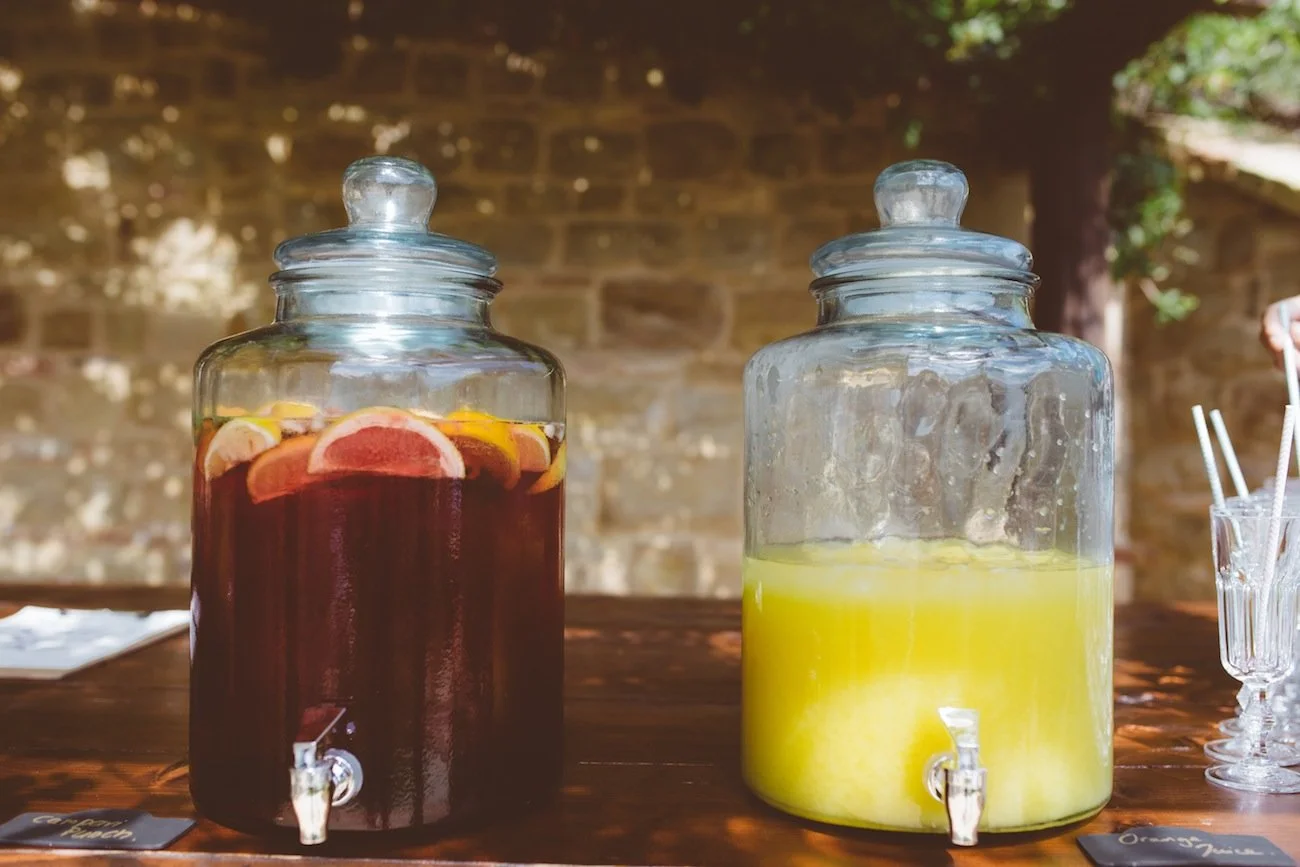 Two large glass beverage dispensers on a wooden table, one filled with dark red liquid with citrus slices, the other with yellow liquid; a stone wall and some green foliage in the background.