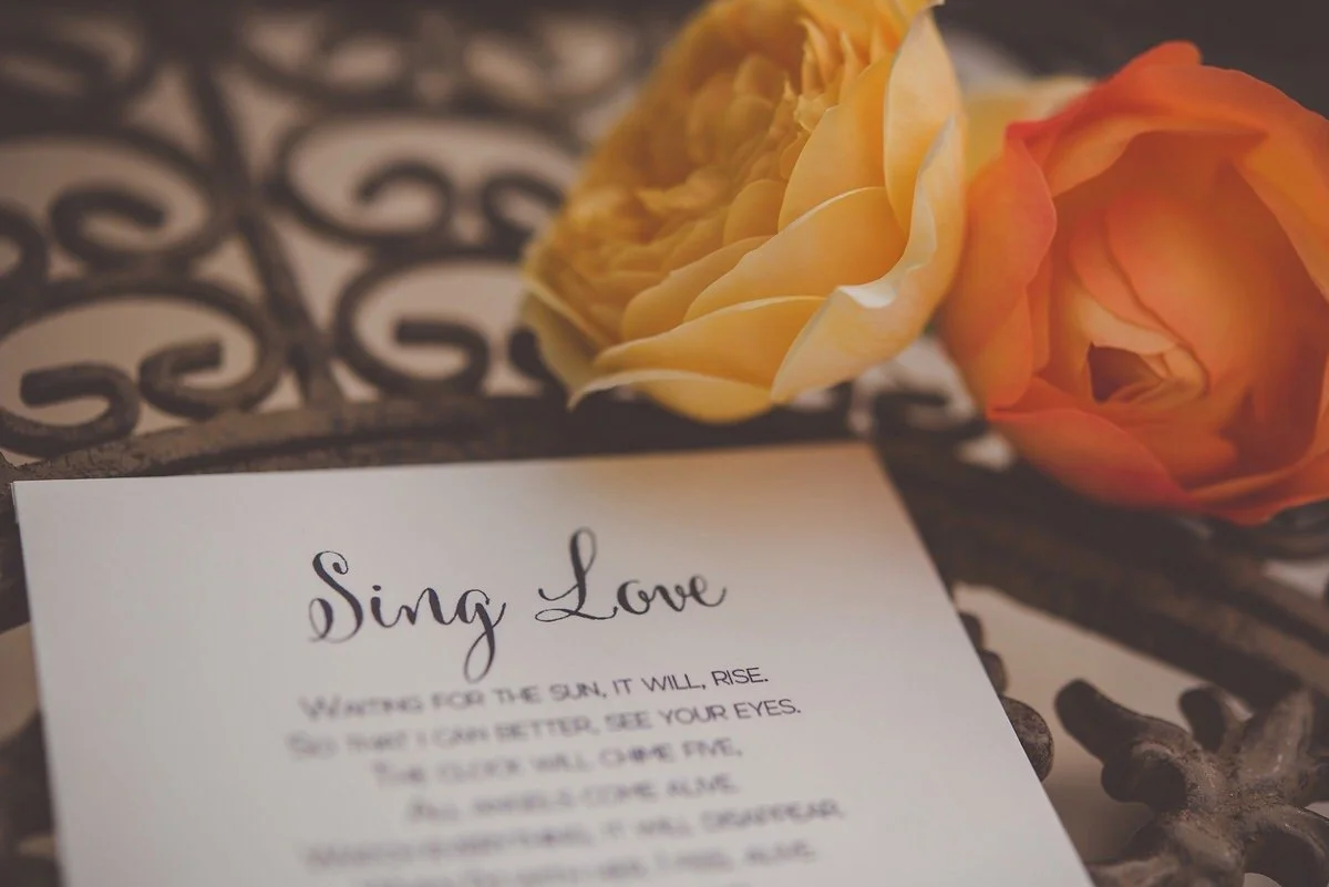 A paper with the title 'Sing Love' and a love poem, placed on a decorative metal surface with two roses, one peach and one orange, resting beside it.