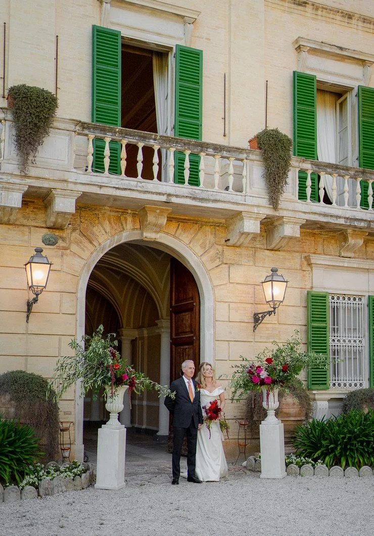 A bride and groom standing outside a historic building with green shutters, floral decorations, and vintage lanterns.