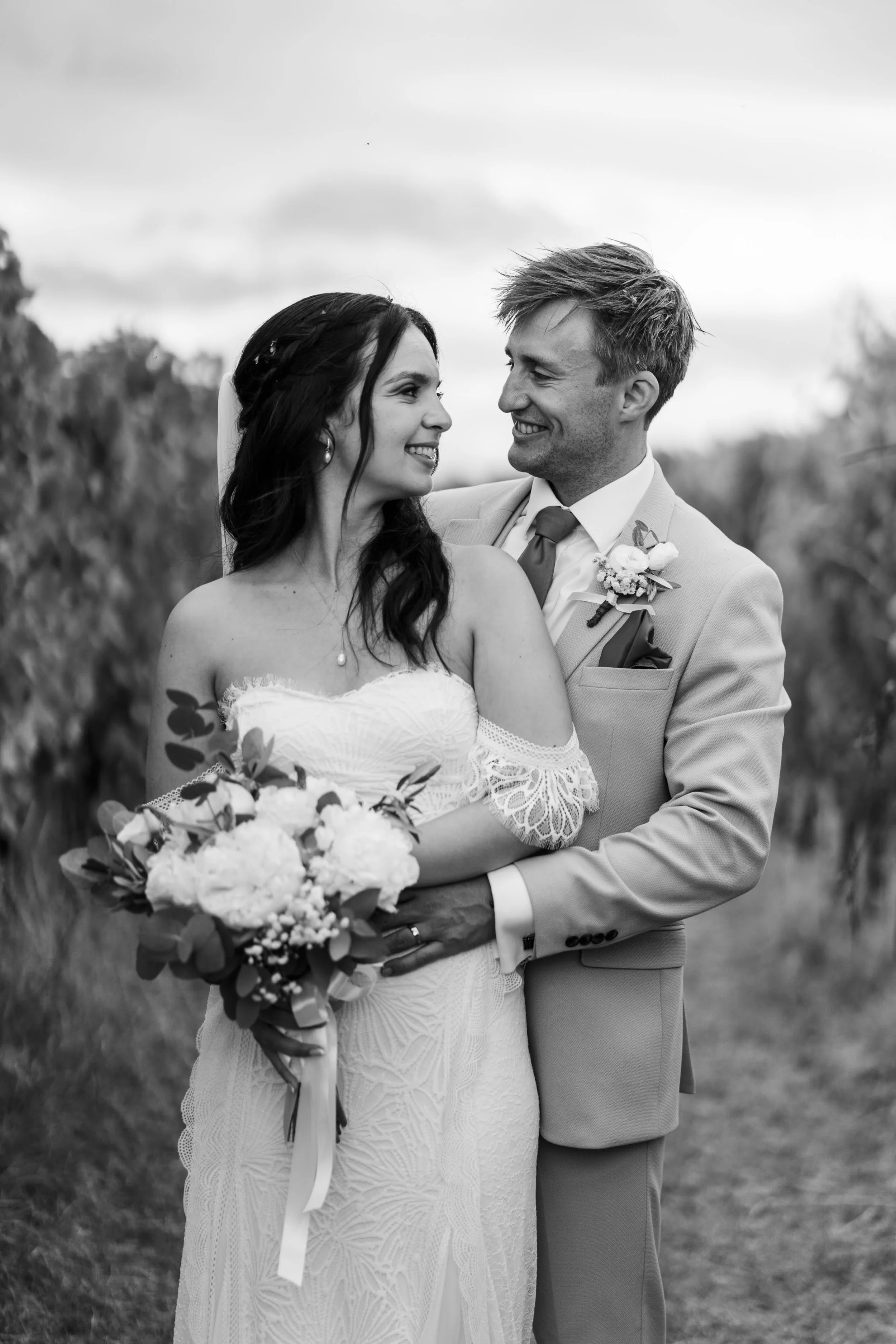 Black and white photo of a wedding couple outdoors, the woman holding a bouquet of flowers and the man smiling at her, both dressed in wedding attire.