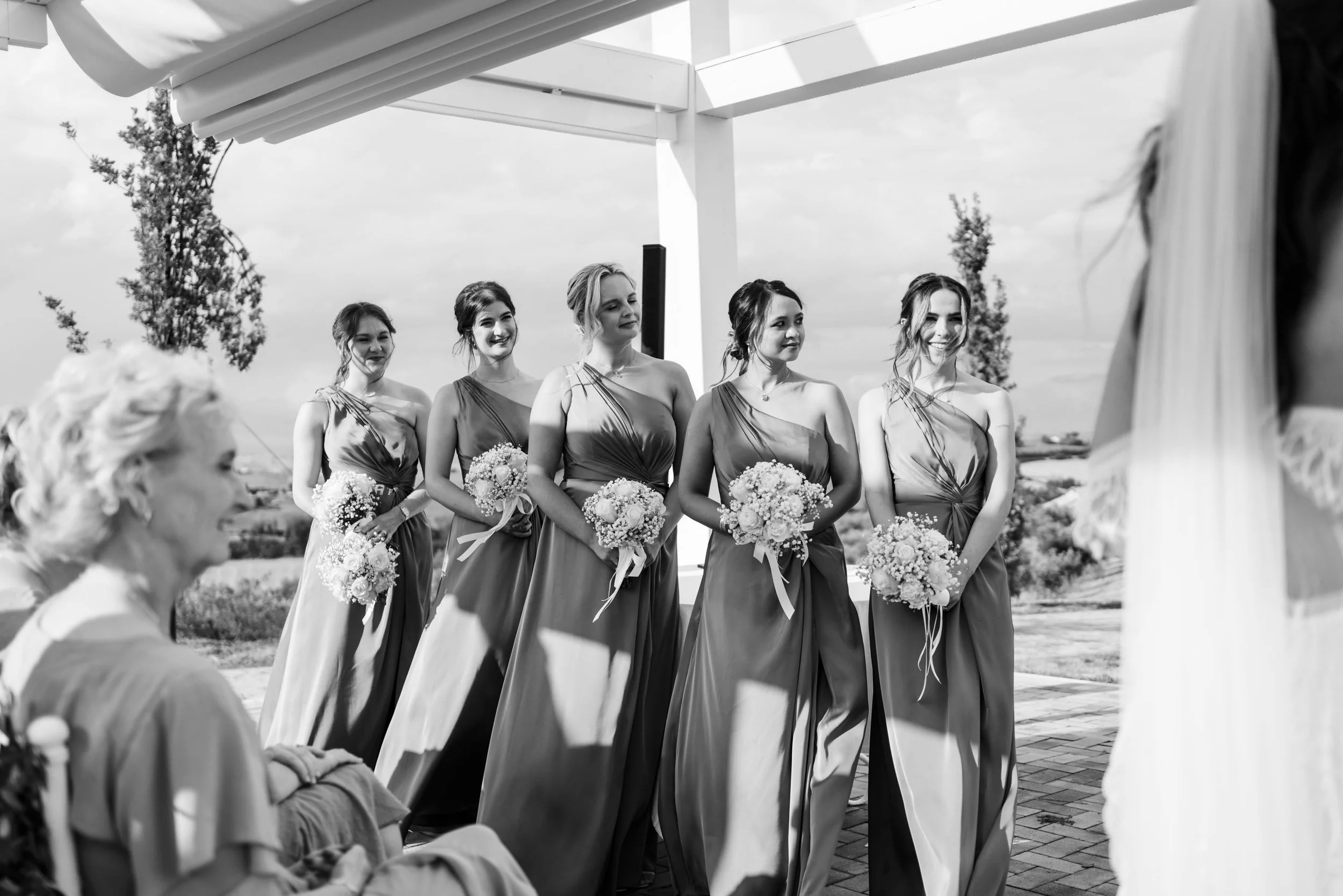 Black and white photo of a bride and her five bridesmaids during a wedding ceremony outdoors, with the bride on the right side partially visible and the bridesmaids standing in a row, holding bouquets, under a white pergola.