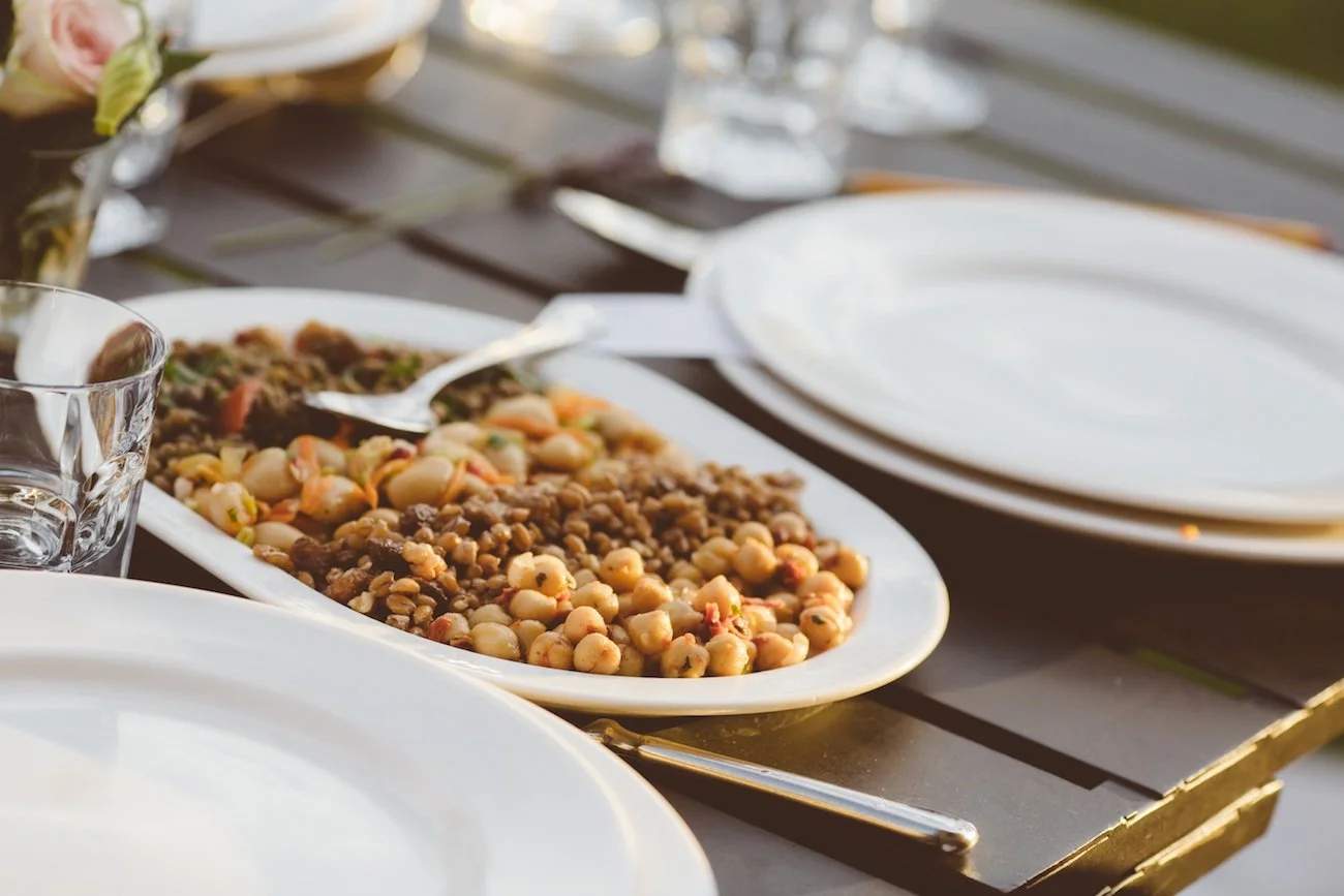 A serving platter of baked beans and chickpeas, with a partially visible white plate and empty glass on a dark wooden table set for a meal.