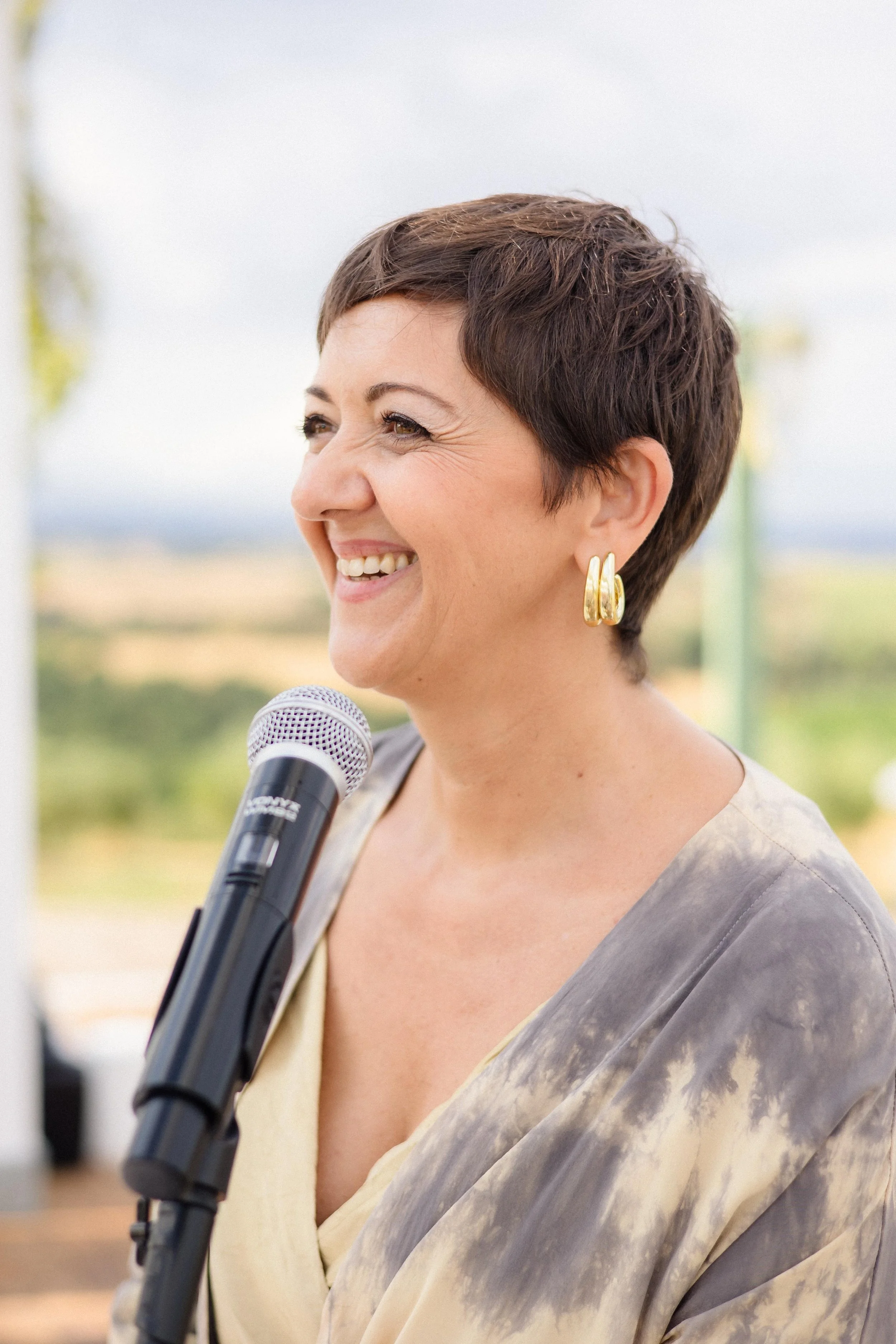 A woman with short dark hair and gold hoop earrings smiling while speaking into a microphone outdoors.