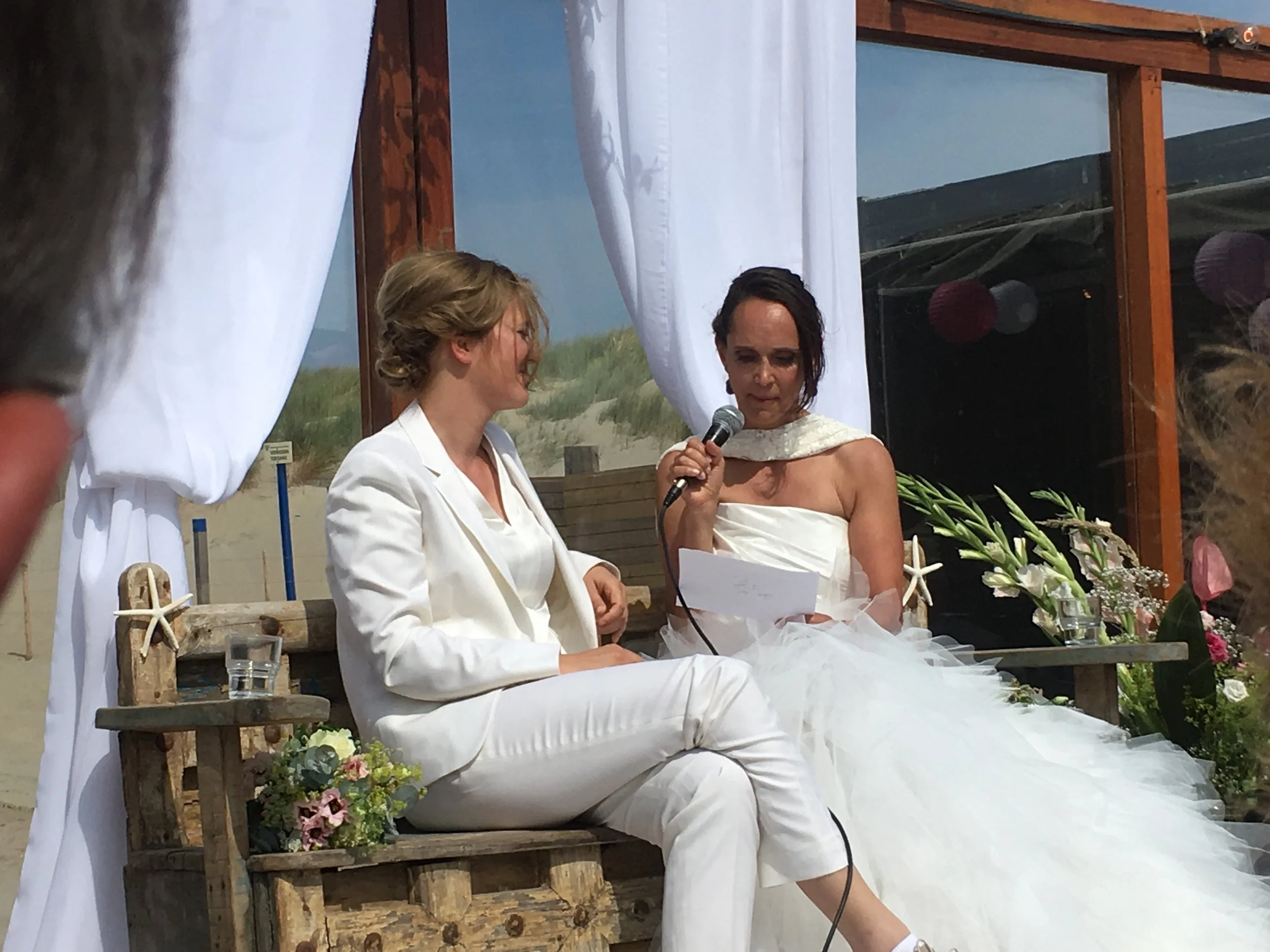 A woman in a white wedding dress is reading vows at a wedding ceremony, with another woman in a white suit sitting beside her, on a beachside setting with white drapes and floral decorations.