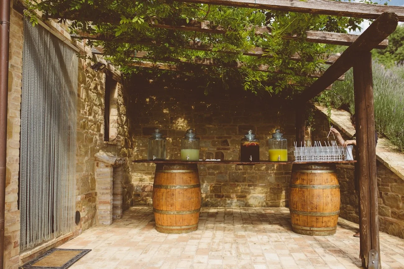 A rustic outdoor drink bar with two large wooden barrels supporting a wooden plank serving as a bar counter, topped with glass jars containing colorful beverages, situated against a stone wall with a canopy of green leaves overhead.