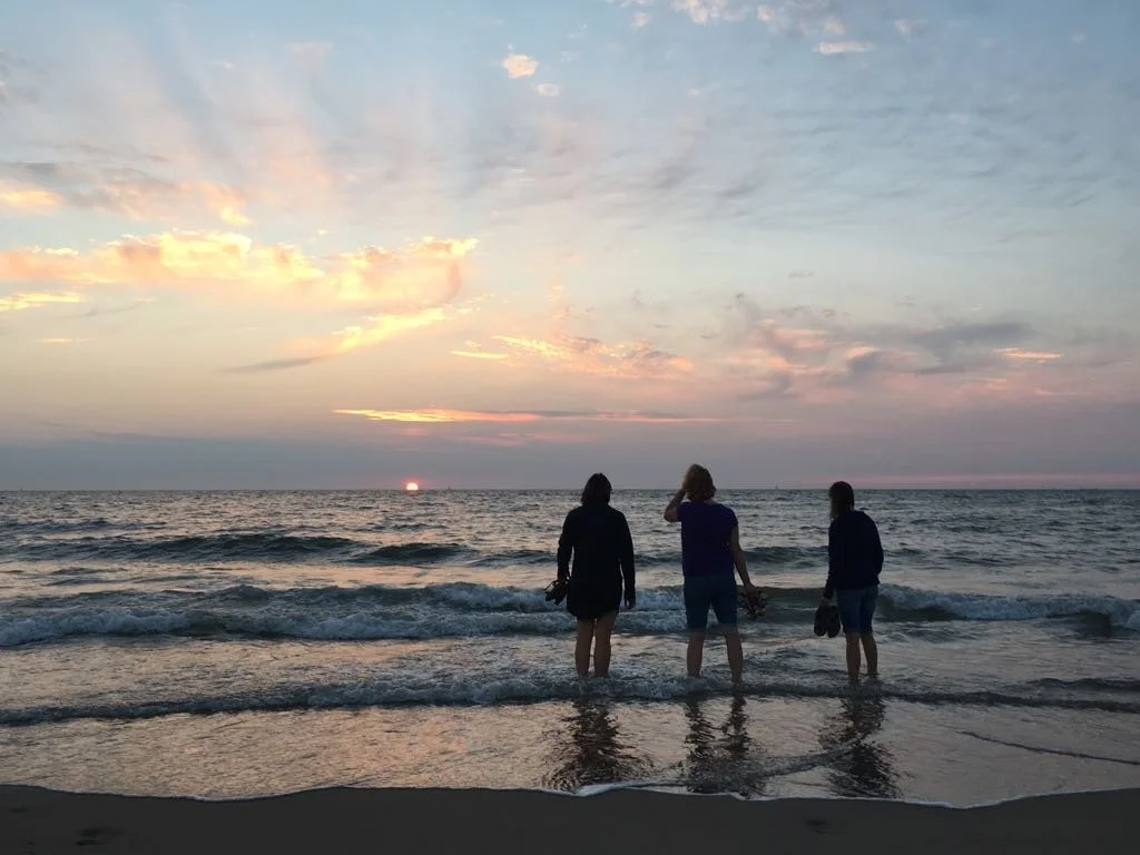 Three people stand in shallow water at the beach during sunset, looking at the horizon with the sun setting over the ocean and pink and orange clouds in the sky.