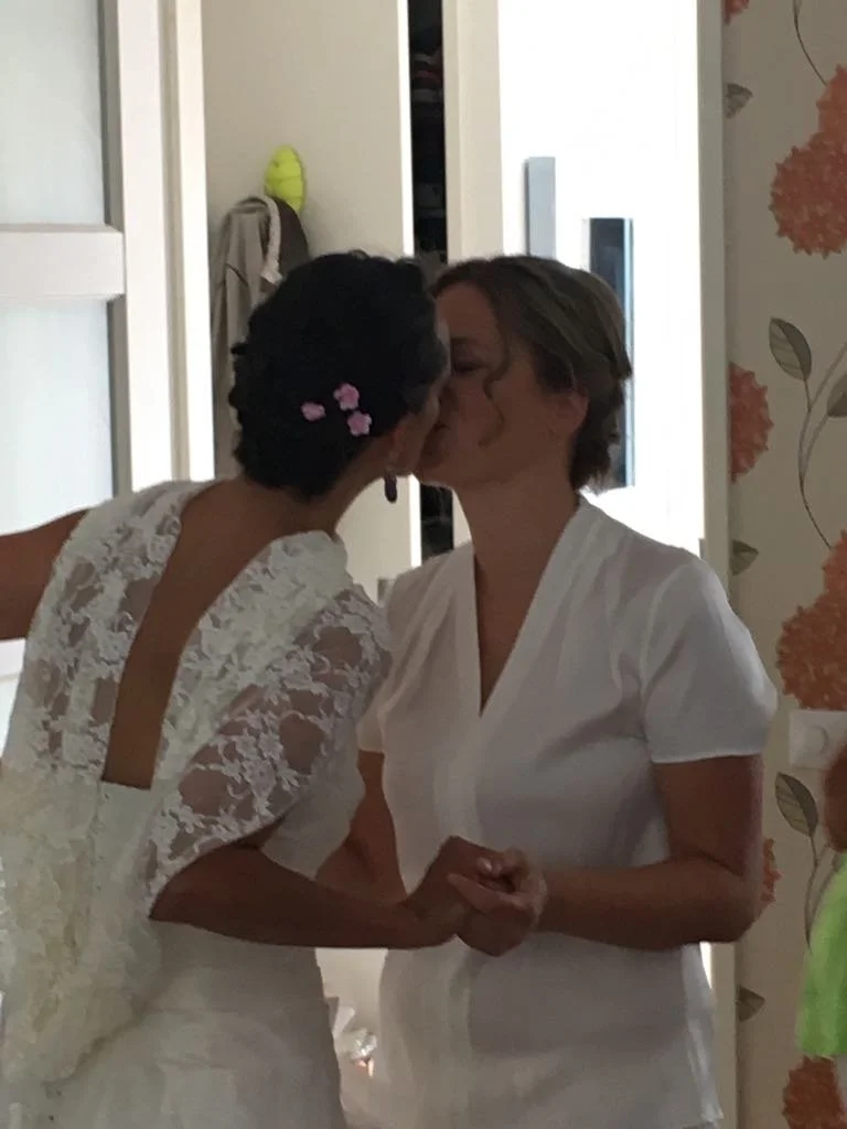Two women in wedding attire sharing a kiss and holding hands indoors, with floral wallpaper and closet doors in the background.