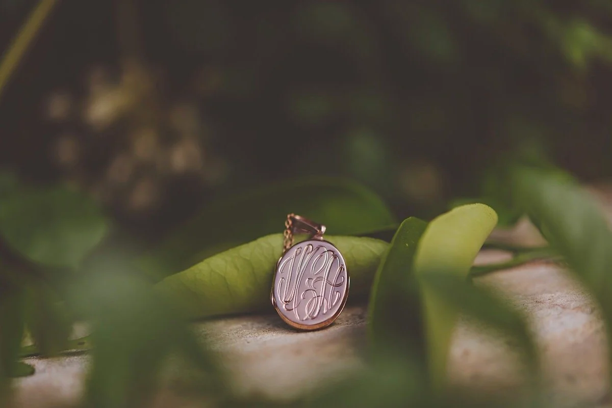 A close-up of a gold pendant with engraved initials resting on a leaf amidst greenery.