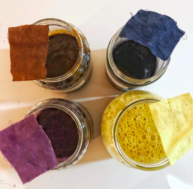 Four glass jars containing different colored liquids, each covered with a piece of fabric, are arranged on a white surface. The liquids are dark brown, black, dark purple, and yellow.