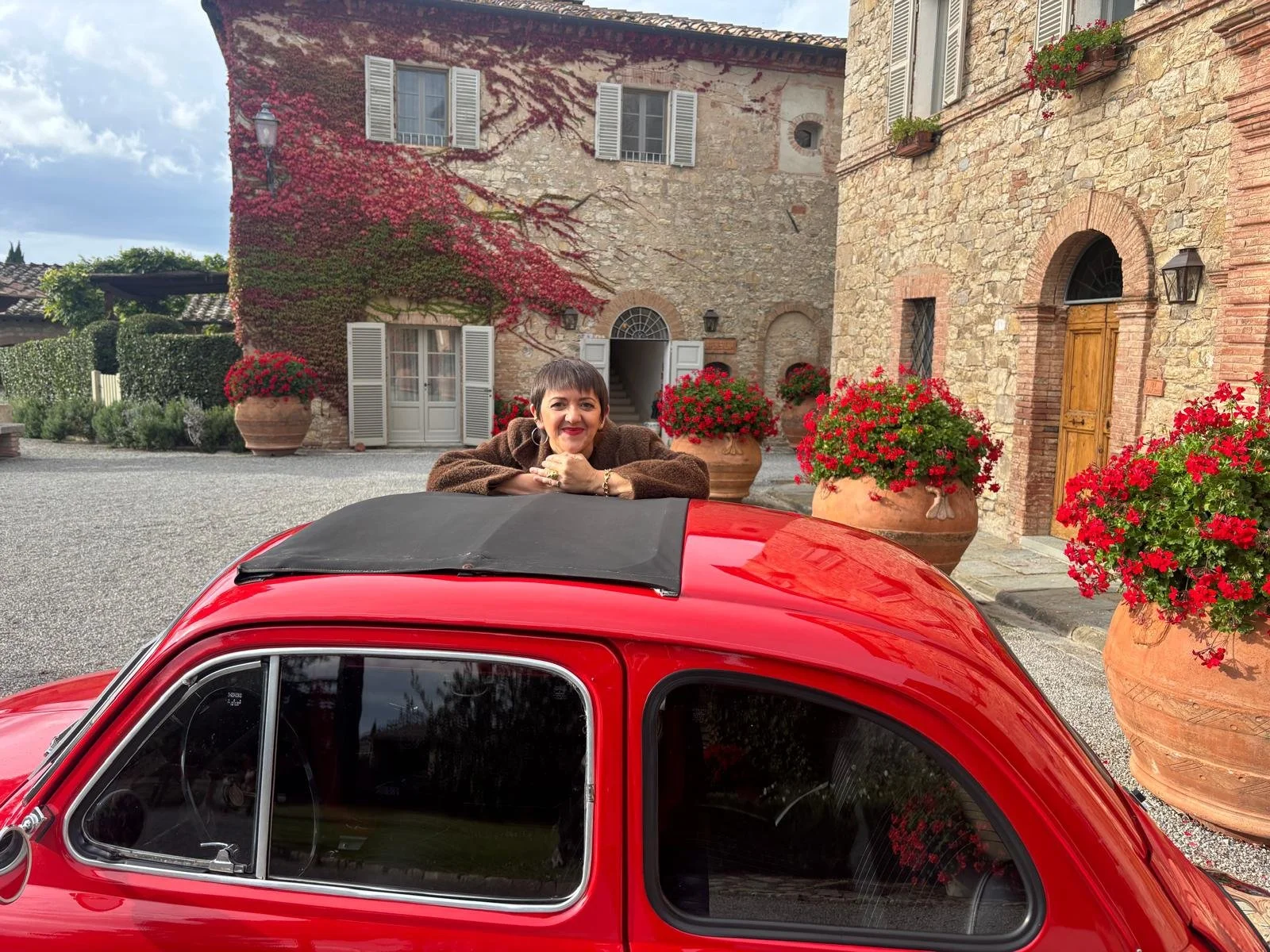Woman leaning on the roof of a red vintage car, smiling, with a European stone building and potted red flowers in the background.