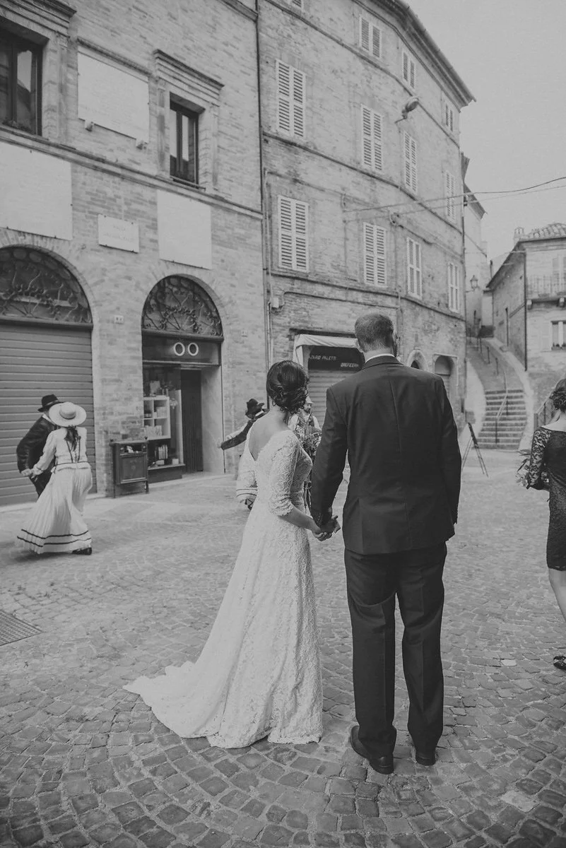 A black-and-white photo of a bride and groom holding hands on a cobblestone street in an old European city, with a building that has shuttered windows and stairs in the background. Other people are visible around them, some dressed in vintage clothin