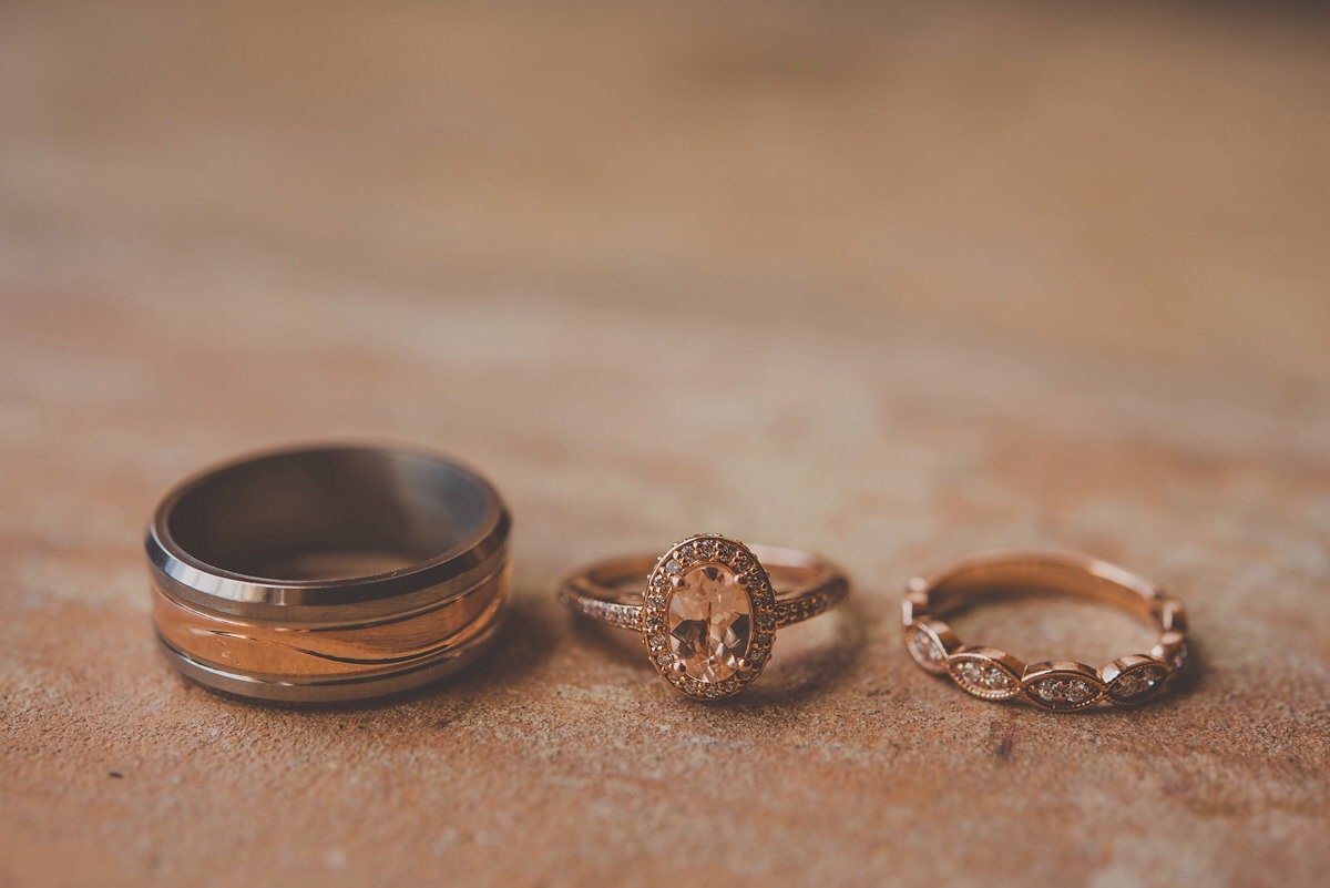 Three rings placed on a textured surface, including a modern wide band in metallic tones, a rose gold oval-shaped gemstone ring with a halo of smaller stones, and a rose gold band with small marquise-shaped stones.