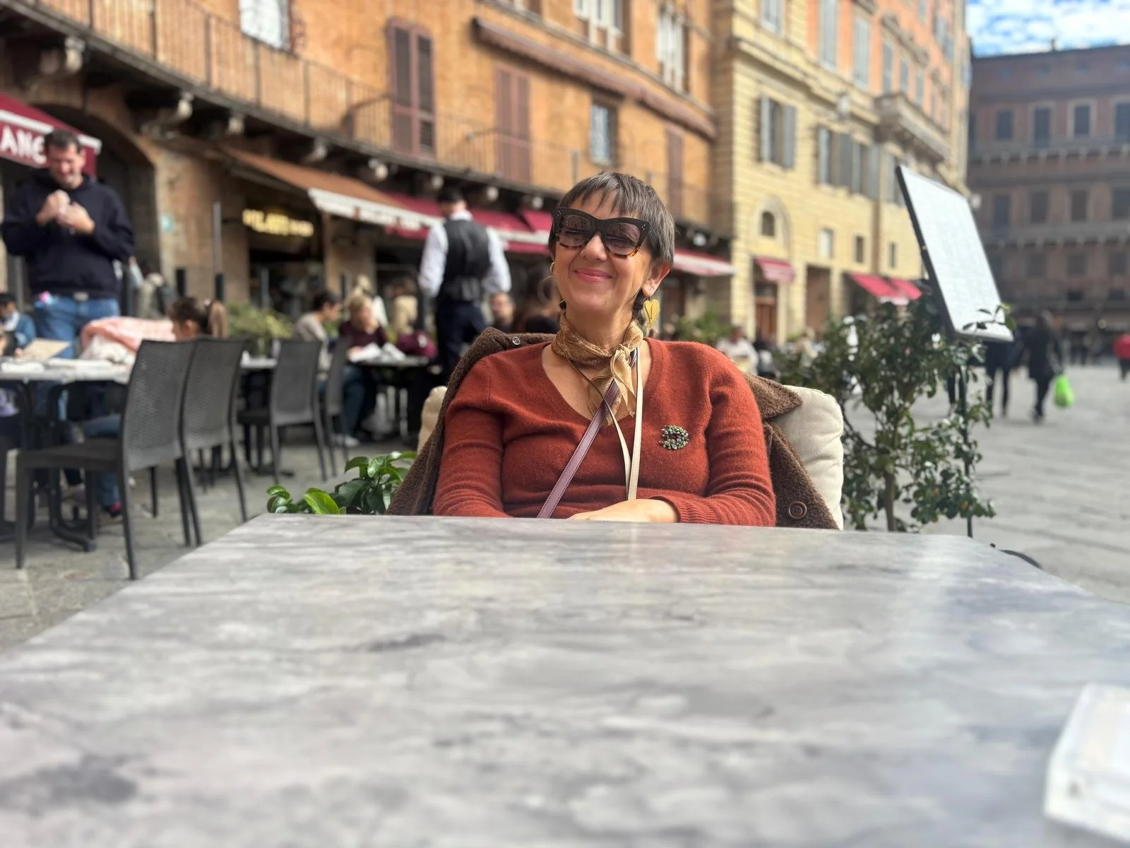 A woman sitting at an outdoor cafe table, smiling, wearing sunglasses, a brown jacket, and a patterned scarf, with a busy city street in the background.