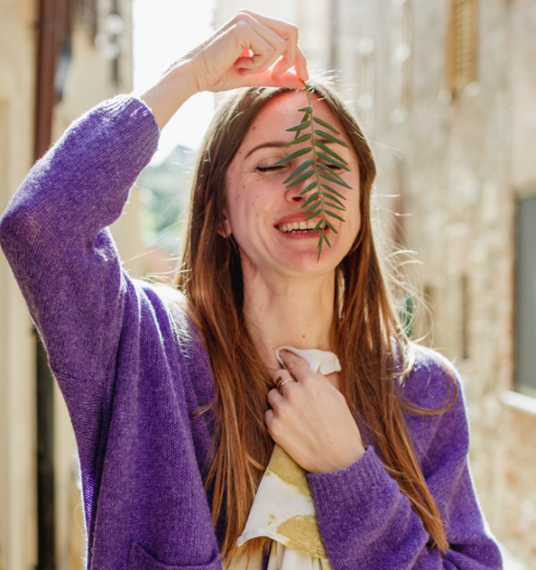 A woman wearing a purple sweater holding a green leaf in front of her face indoors, smiling.